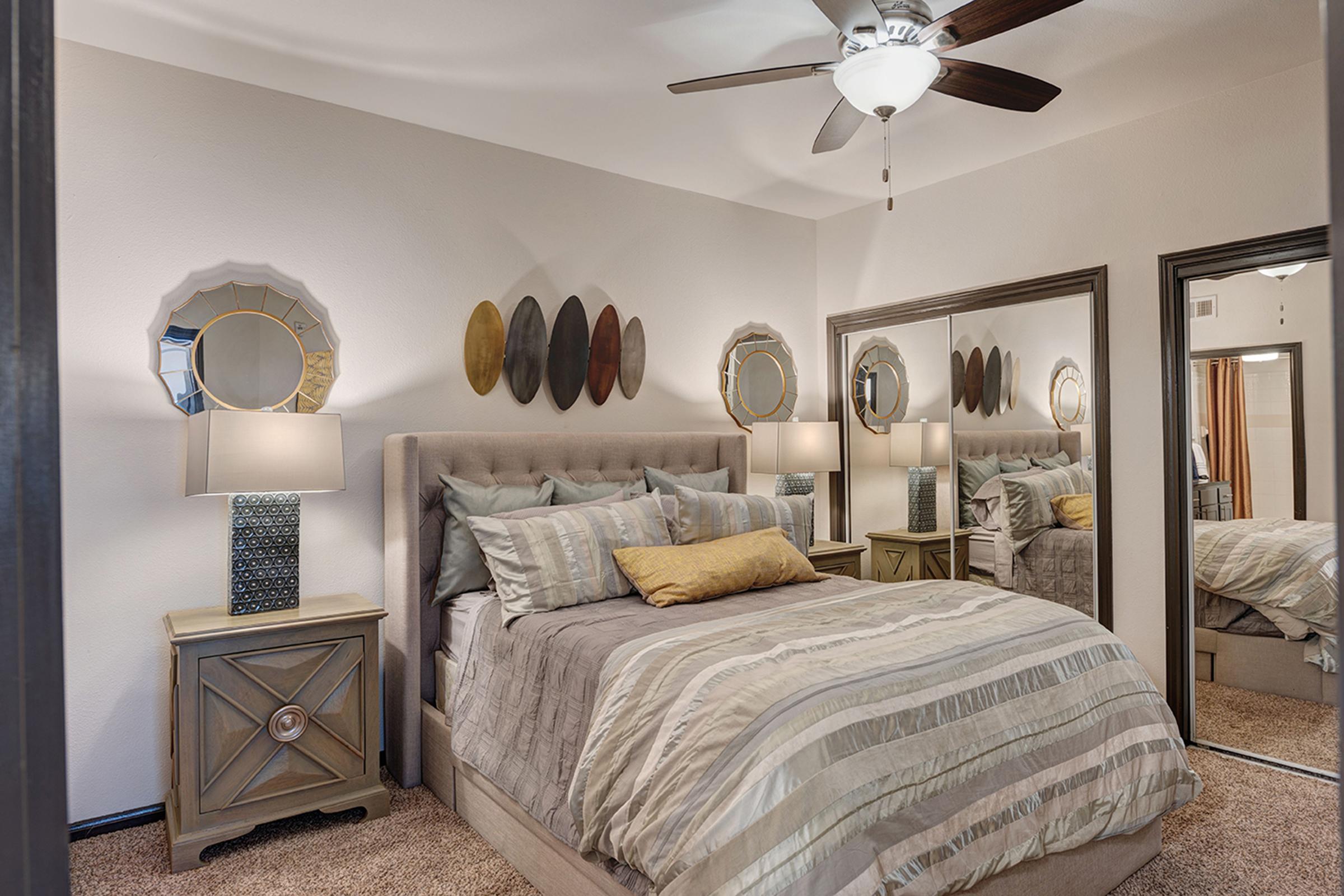 A cozy bedroom featuring a queen-sized bed with striped bedding, decorative pillows, and a soft throw blanket. There are bedside tables with lamps, a large mirror, and an accent wall with decorative plates. A ceiling fan adds to the ambiance, and the room has warm, inviting tones.