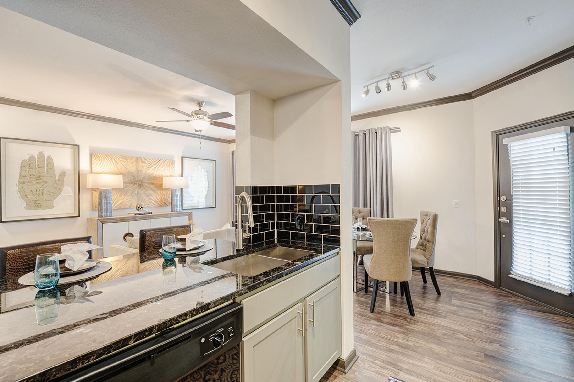 A modern apartment interior featuring a kitchen with a granite countertop, a dining area with stylish chairs, and a living room with decorative art and lighting. Natural light floods the space through a door with blinds, enhancing the contemporary decor and warm ambiance.