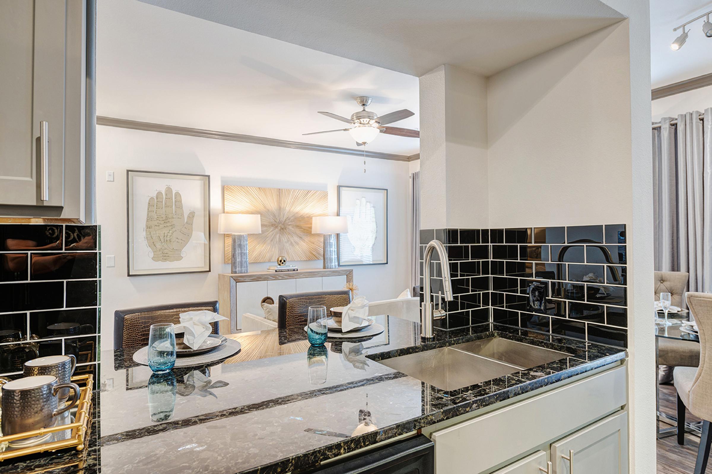 A modern kitchen with a black and white color scheme. It features dark countertops, a stainless steel sink, and a breakfast bar. In the background, a living area is visible with stylish decor, including wall art and lamps. Bright lighting and a ceiling fan add ambiance to the open space.