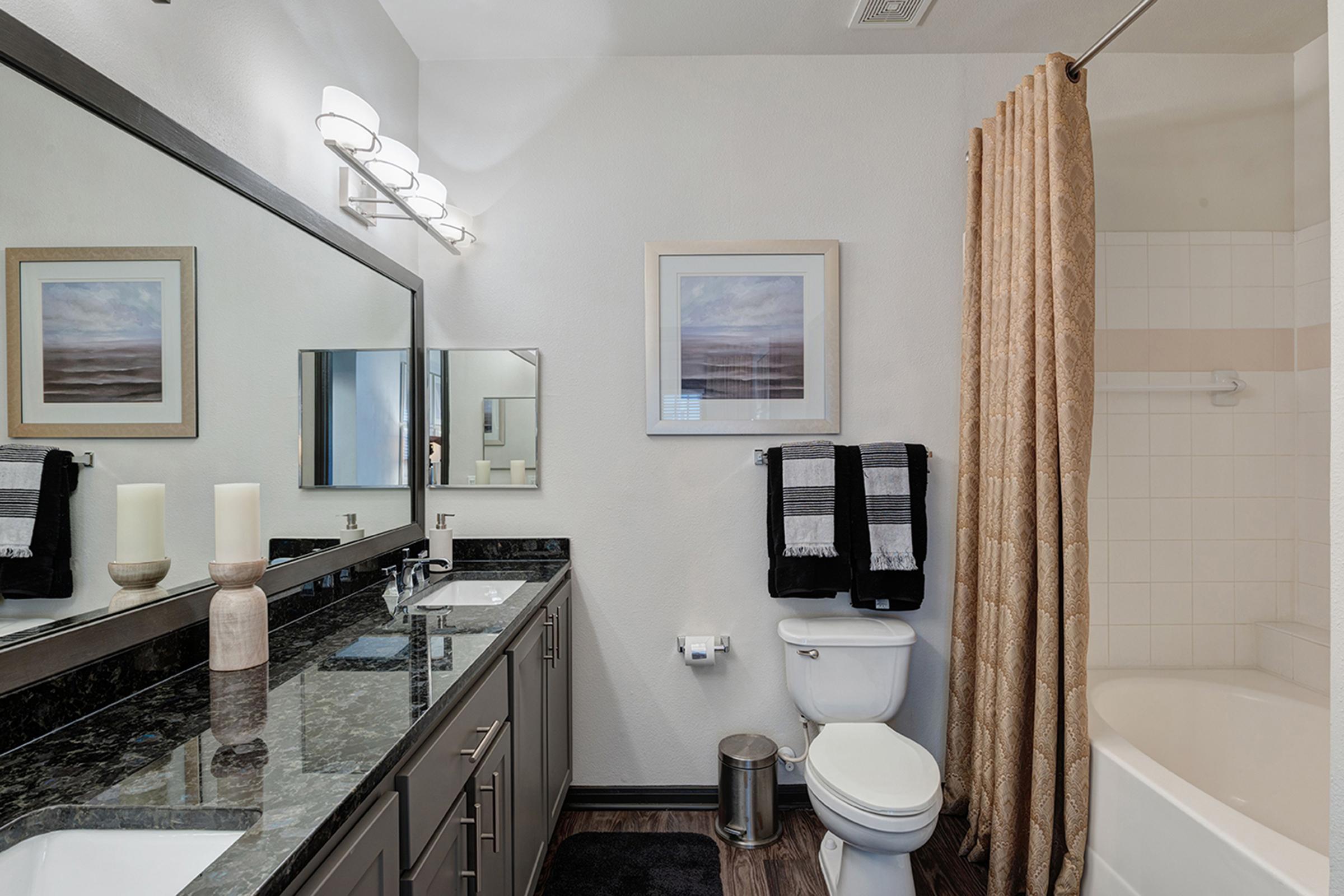 A modern bathroom featuring a granite vanity with a large mirror, decorative candles, and neatly arranged towels. To the right, there's a bathtub with a shower curtain. The walls are painted a light color, and a framed artwork hangs above the vanity, adding a touch of elegance to the space.