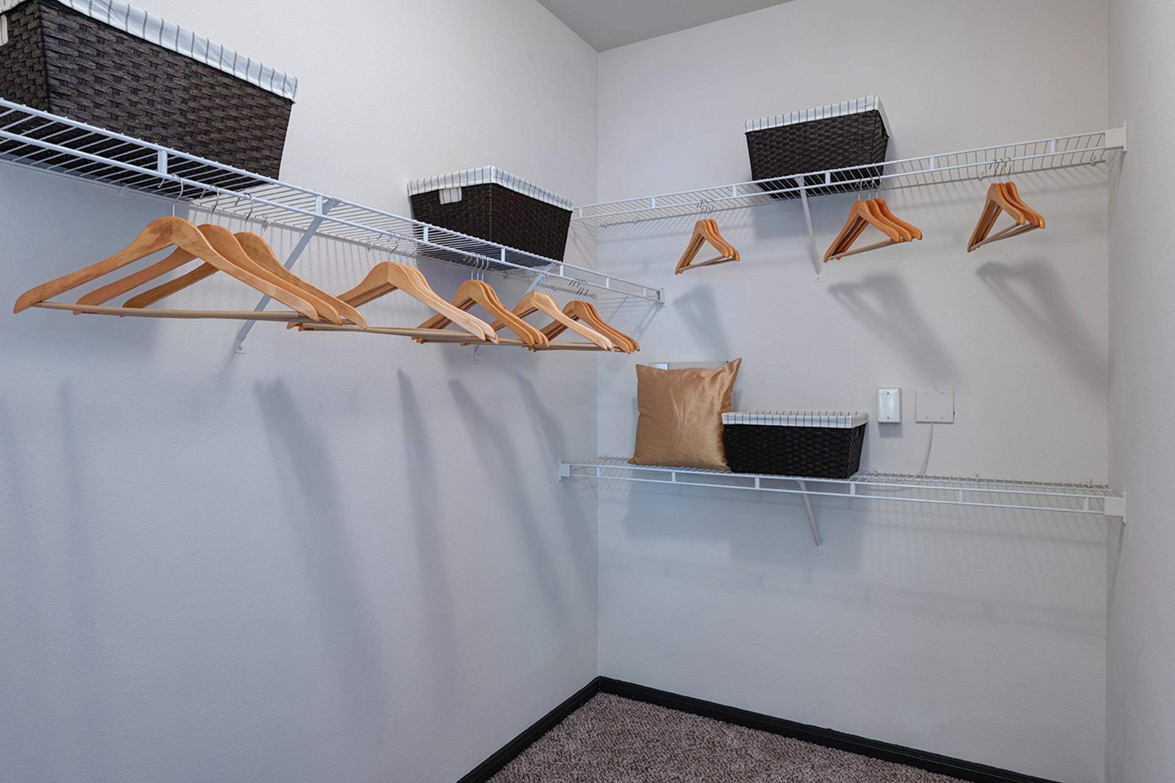 A well-organized closet featuring wire shelves with several wooden hangers, two woven storage baskets, and a decorative pillow. The walls are painted white, and the floor is carpeted, creating a clean and minimalist appearance.
