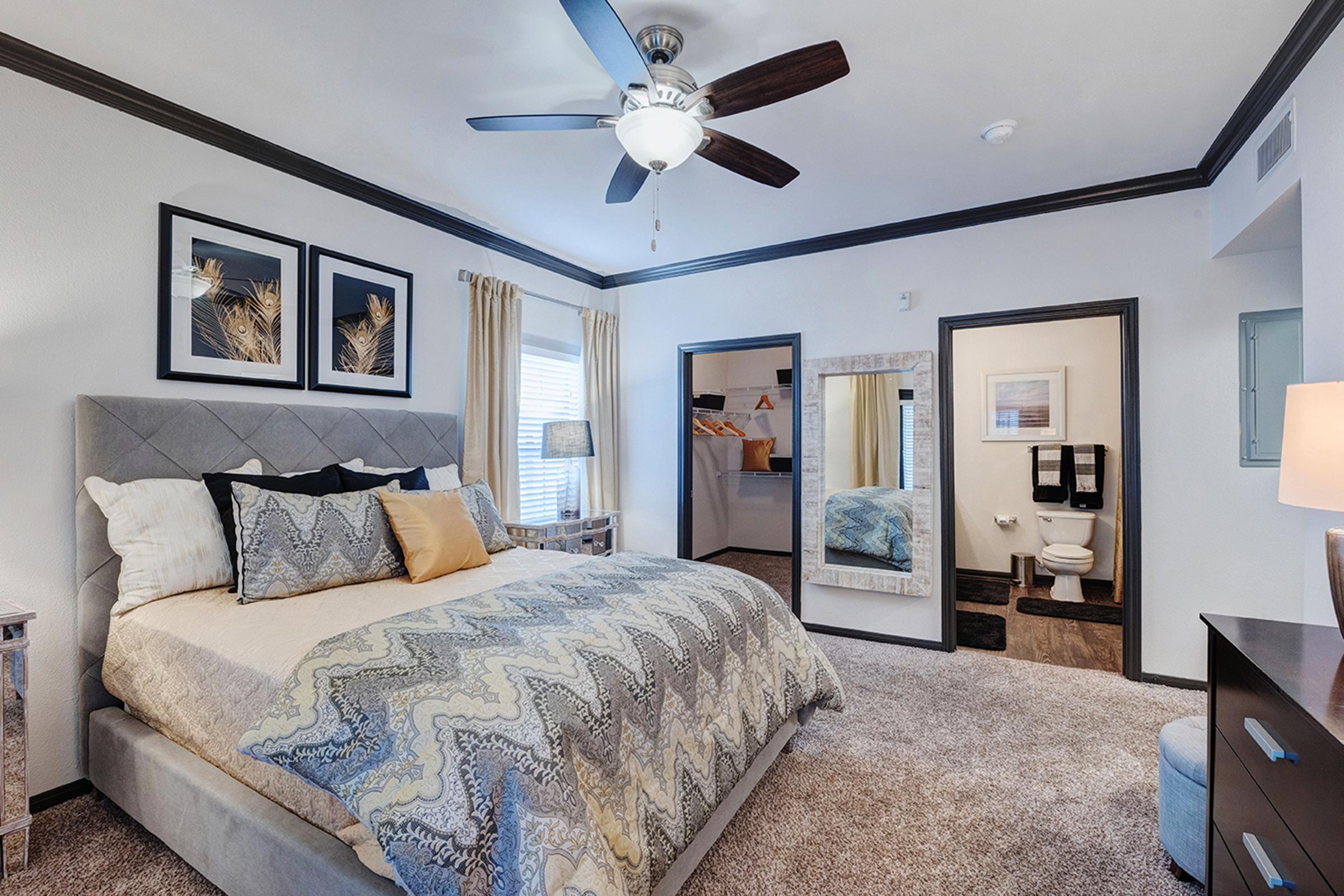 A cozy bedroom featuring a large bed with decorative pillows and a stylish quilt. There are two framed artworks above the bed, a ceiling fan, and a large mirror reflecting the room. Natural light comes through a window with sheer curtains, and a bathroom is visible in the background. The overall decor is modern and inviting.