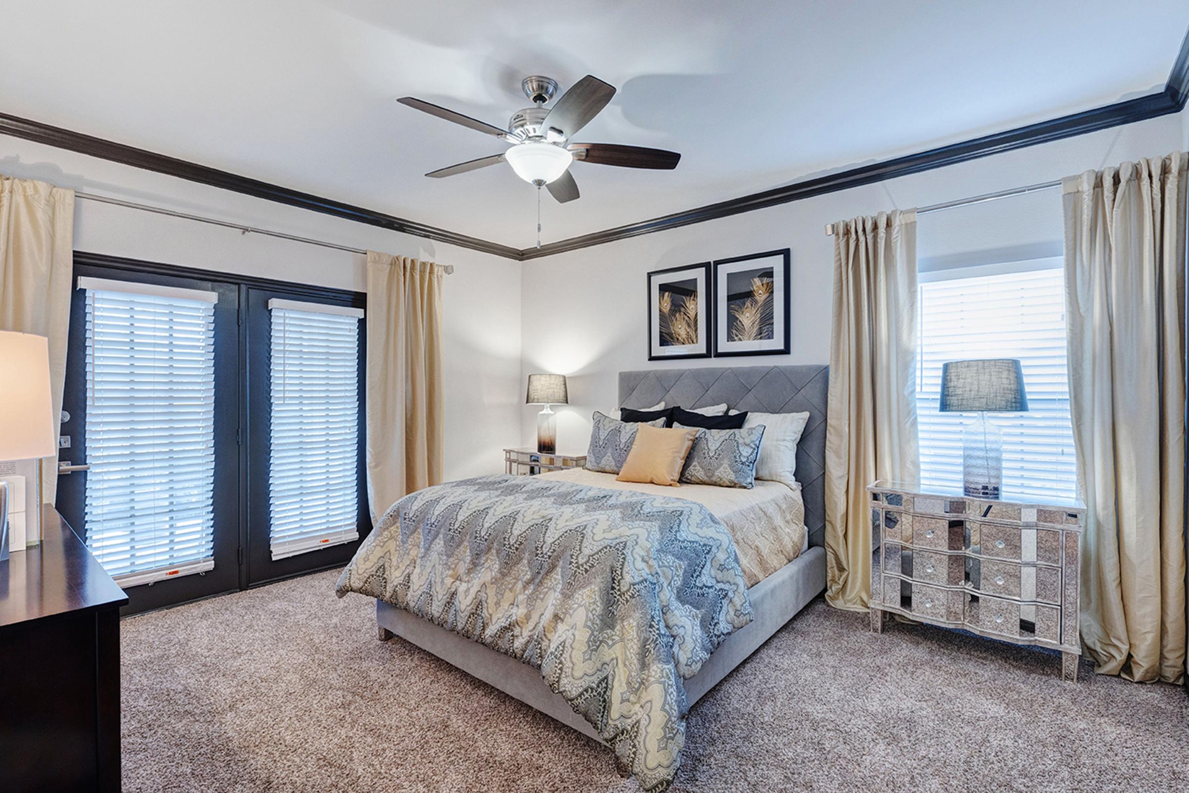 A beautifully decorated bedroom featuring a plush king-sized bed with a patterned comforter, two bedside lamps, and elegant curtains. The room has dark wood accents, a ceiling fan, and art decor on the walls. Large windows with shutters allow natural light to fill the space, creating a cozy atmosphere.