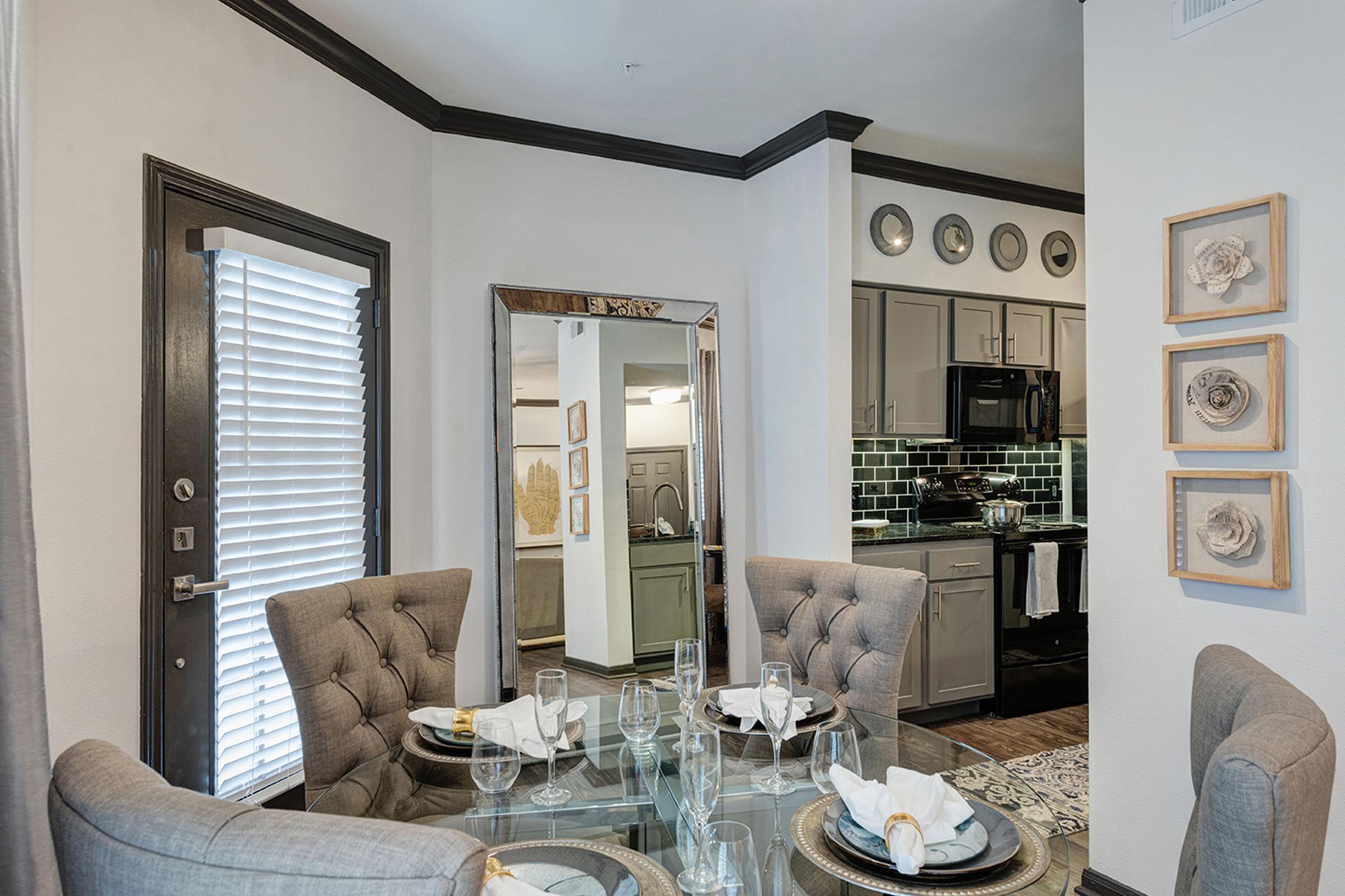 A stylish dining area with a glass table set for a meal, featuring elegant tableware and decorative napkins. The walls are adorned with framed art, and a large mirror reflects the space. In the background, there’s a modern kitchen with dark cabinetry and decorative plates displayed on the wall. Bright natural light streams in through a window.