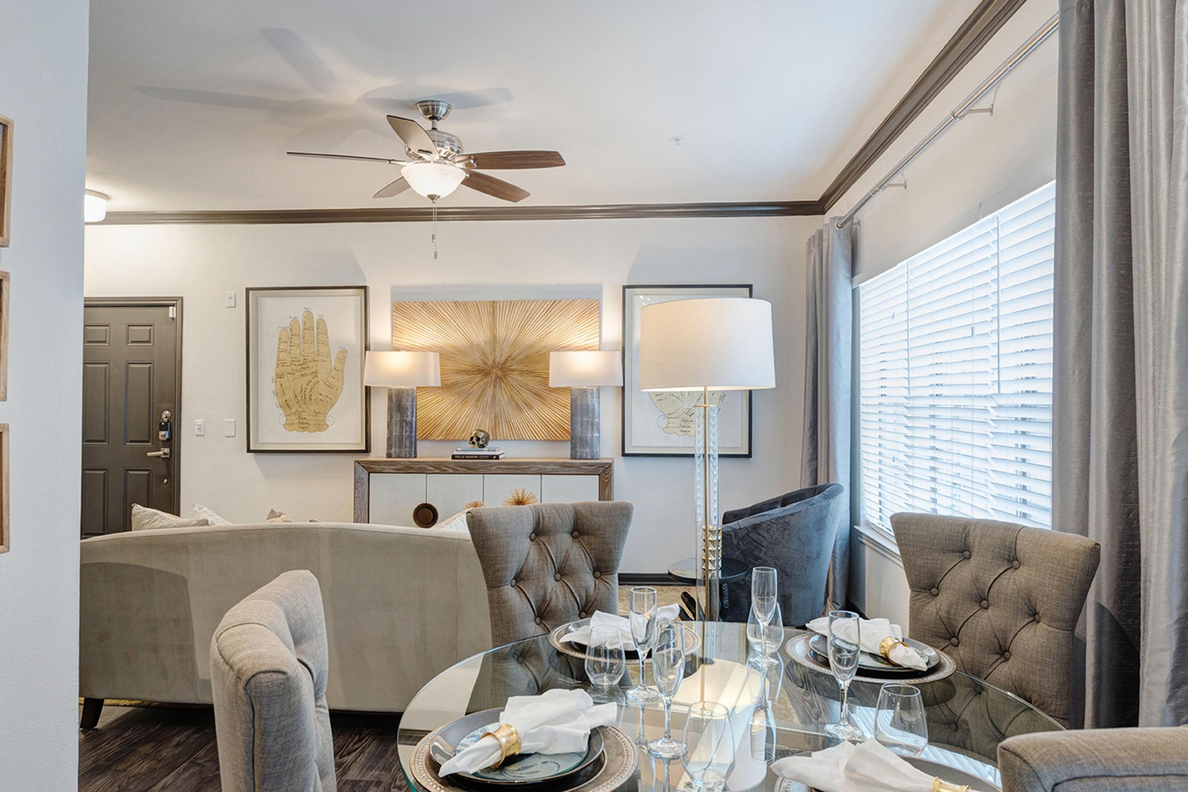 A modern dining area featuring a glass table set with elegant tableware. Surrounding the table are tufted chairs. The room includes a stylish console table, decorative artwork on the walls, a ceiling fan, and large windows with sheer curtains letting in natural light. Overall, the décor is contemporary and inviting.