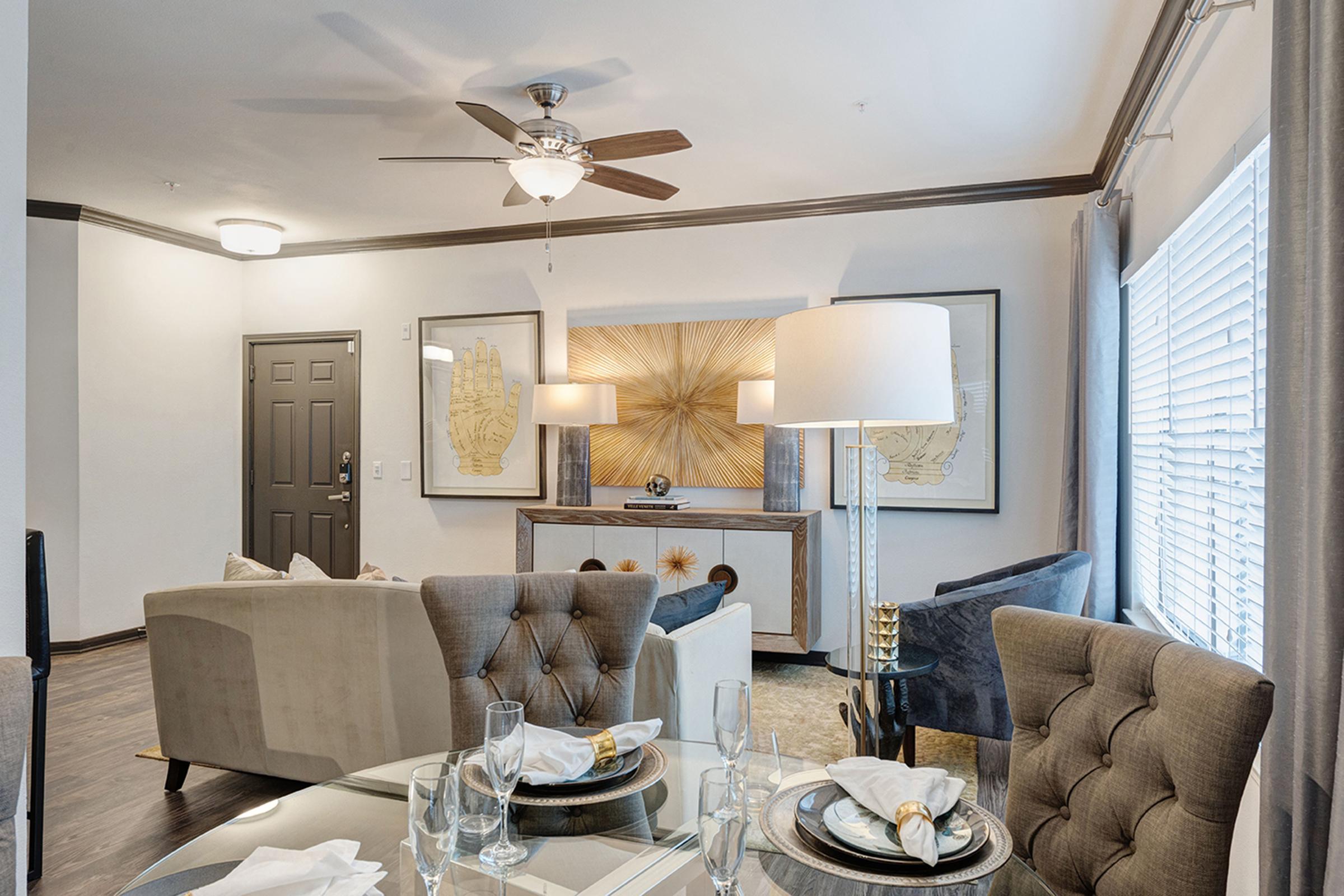 A modern living area featuring a stylish gray sofa with tufted details, a round dining table set for two with elegant tableware, and a decorative lamp. The walls are adorned with framed artwork, and natural light enters through window blinds, creating a warm and inviting atmosphere.