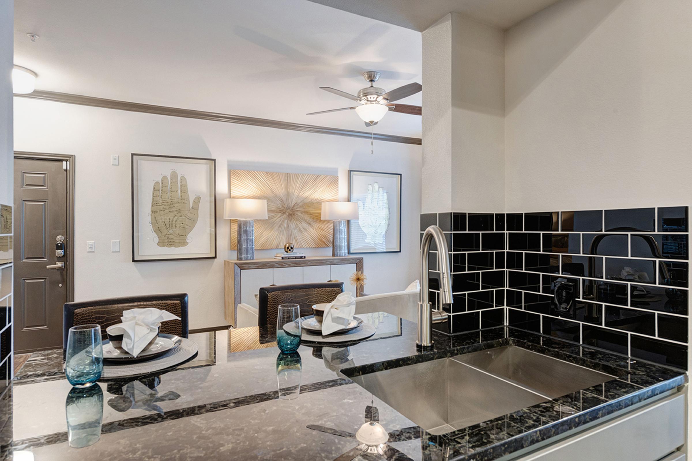 Modern kitchen with black and white tile backsplash, stainless steel sink, and elegant dining setup. The dining table features two plates with decorative napkins and blue glasses, while the background shows light fixtures and wall art, creating a stylish and inviting living space.