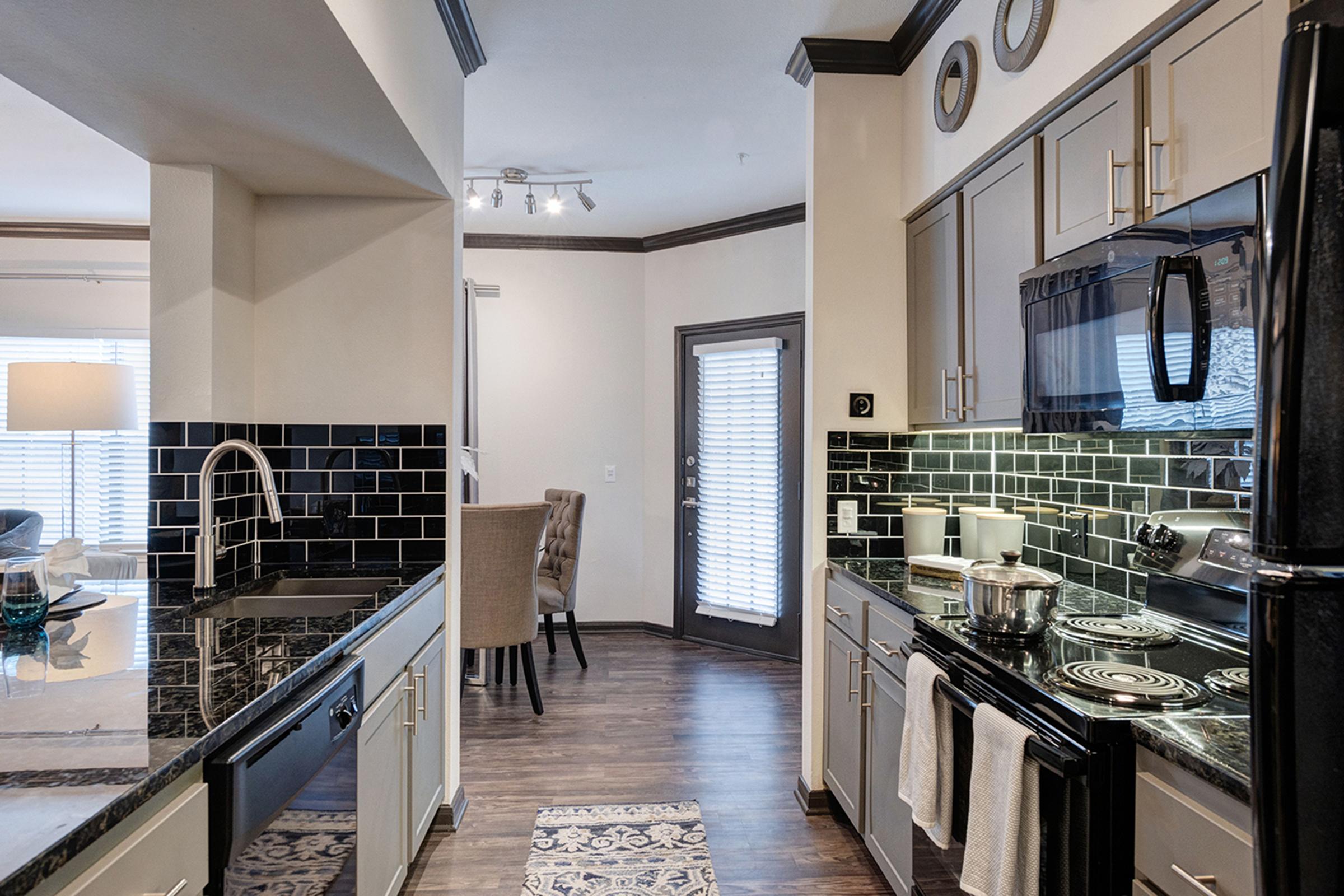 Modern kitchen featuring black and white color scheme, stainless steel appliances, and granite countertops. A dining area with a chair is visible through an open doorway, while natural light fills the space from a nearby window. The design blends functionality with contemporary aesthetics.