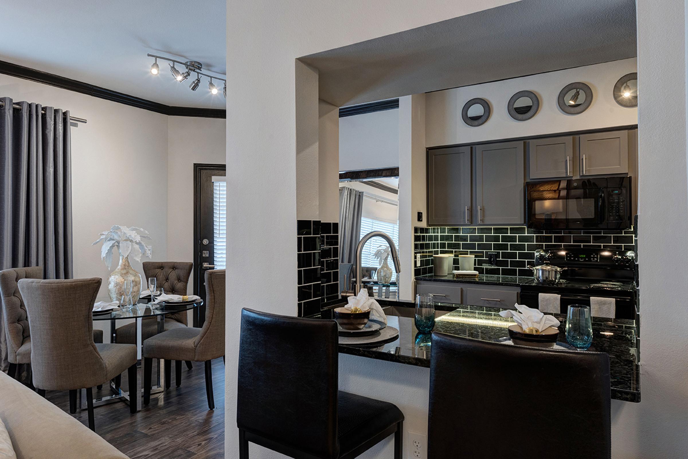 A modern kitchen and dining area featuring dark cabinetry, a stylish black backsplash, and a round glass table with upholstered chairs. The space is well-lit with ceiling lights and has decorative elements like a vase and dinnerware, creating a cozy and inviting atmosphere.