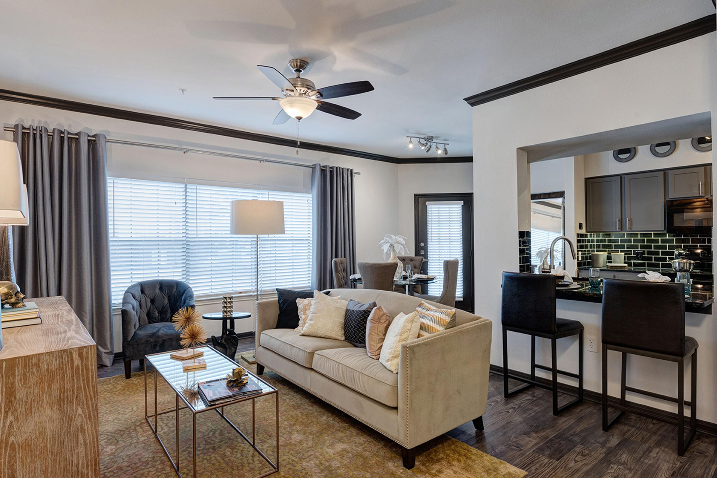 A modern living room featuring a beige sofa with decorative pillows, a stylish coffee table, and a cozy armchair. Natural light streams through large windows dressed with gray curtains. A dining area with a table and chairs is visible, along with a contemporary kitchen in the background, highlighted by dark cabinetry and green tile backsplash.