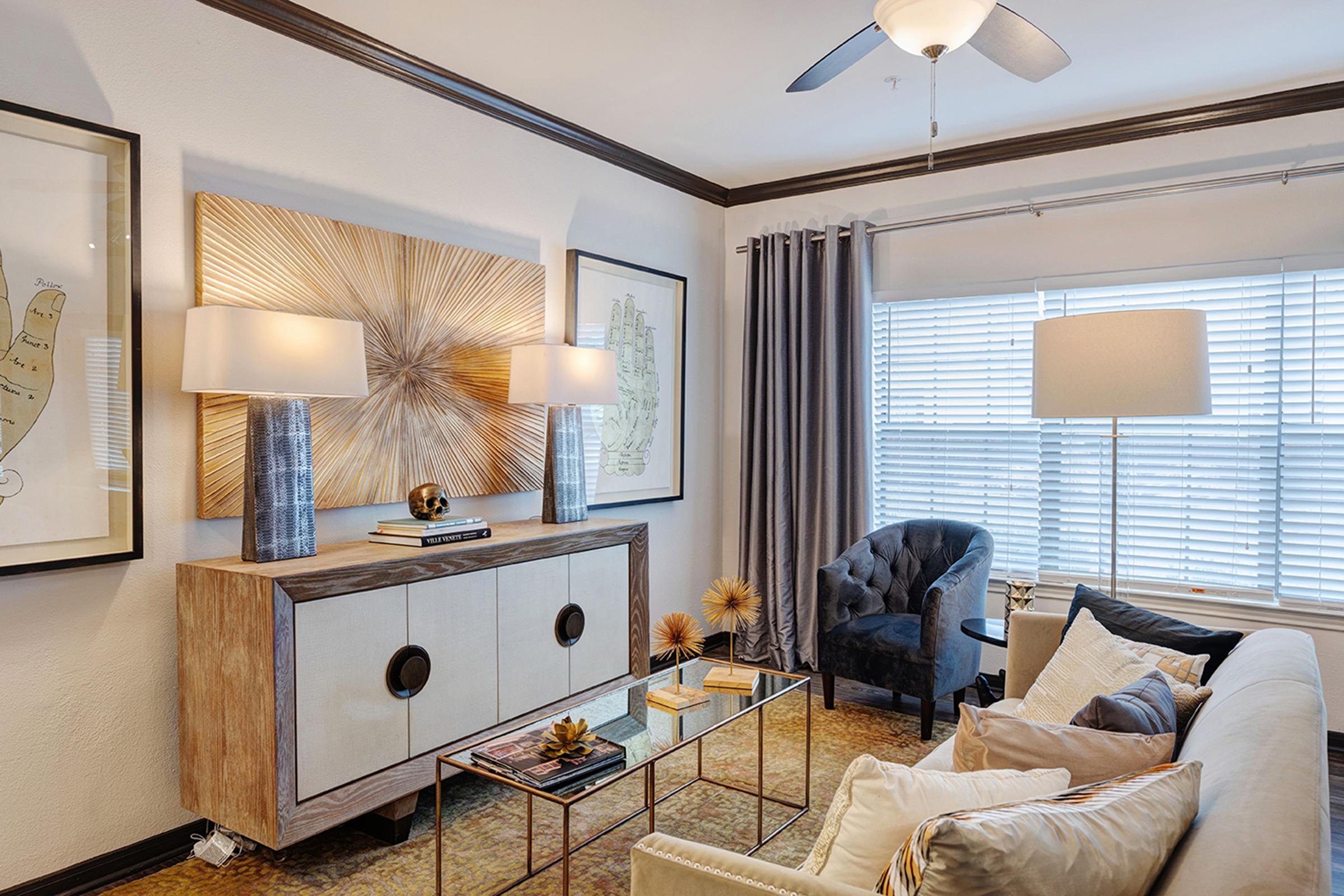 A stylish living room featuring a light-colored sofa with decorative pillows, an elegant glass coffee table, and a wooden console with contemporary lamps. The walls are adorned with framed art, and large windows with sheer curtains allow natural light to fill the space, creating a welcoming atmosphere.
