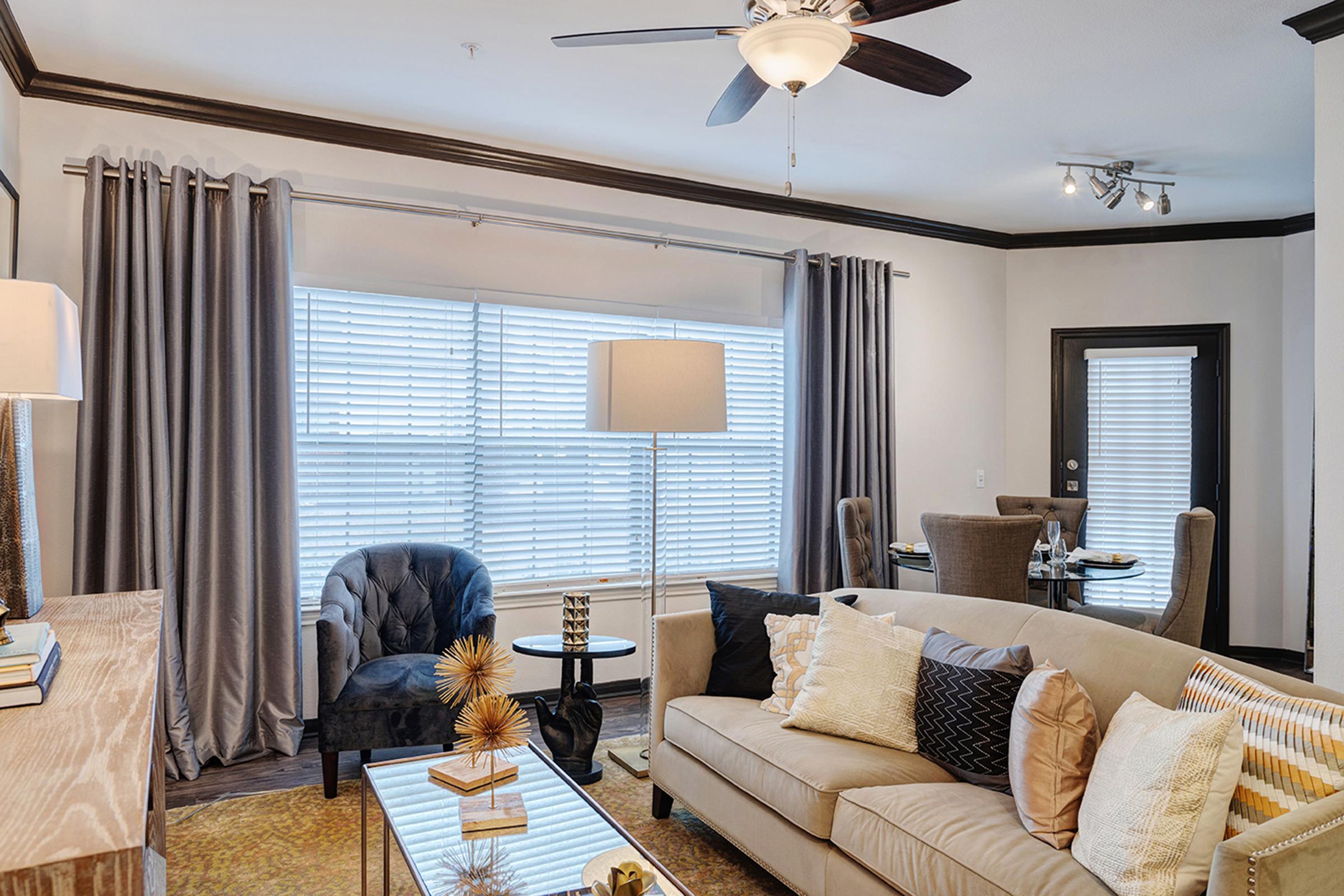 A modern living room featuring a beige sofa with decorative pillows, a glass coffee table, and an artistic centerpiece. Large windows with blinds provide natural light, and the space includes a ceiling fan, a floor lamp, and stylish curtains. A dining area is visible in the background with a glass table and chairs.