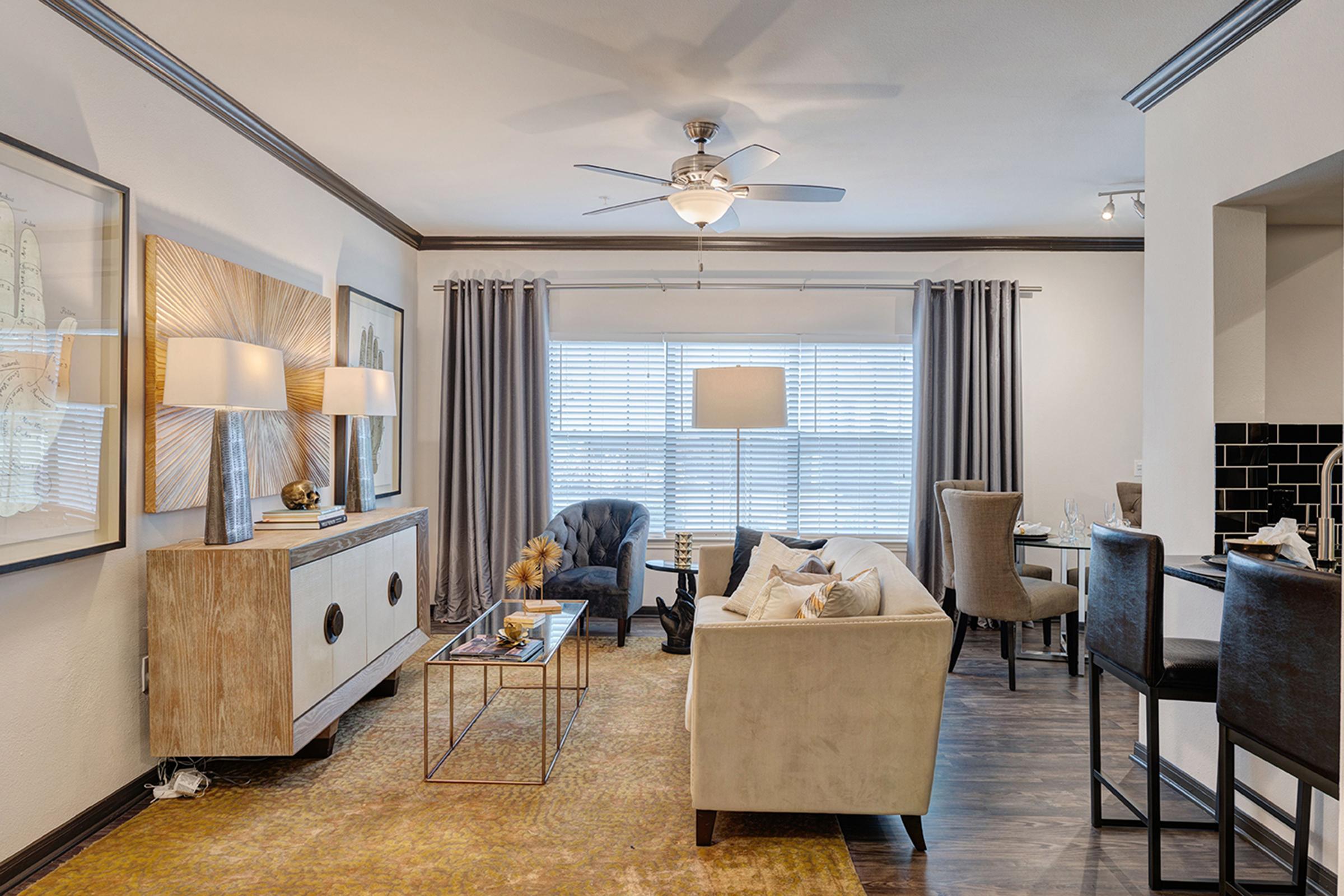 A cozy living room featuring a cream-colored sofa, decorative throw pillows, and a coffee table. There's a sleek sideboard with lamps and artwork on the walls. Natural light floods in through window drapes, and a dining area with high chairs is visible in the background. The decor combines modern and inviting elements.