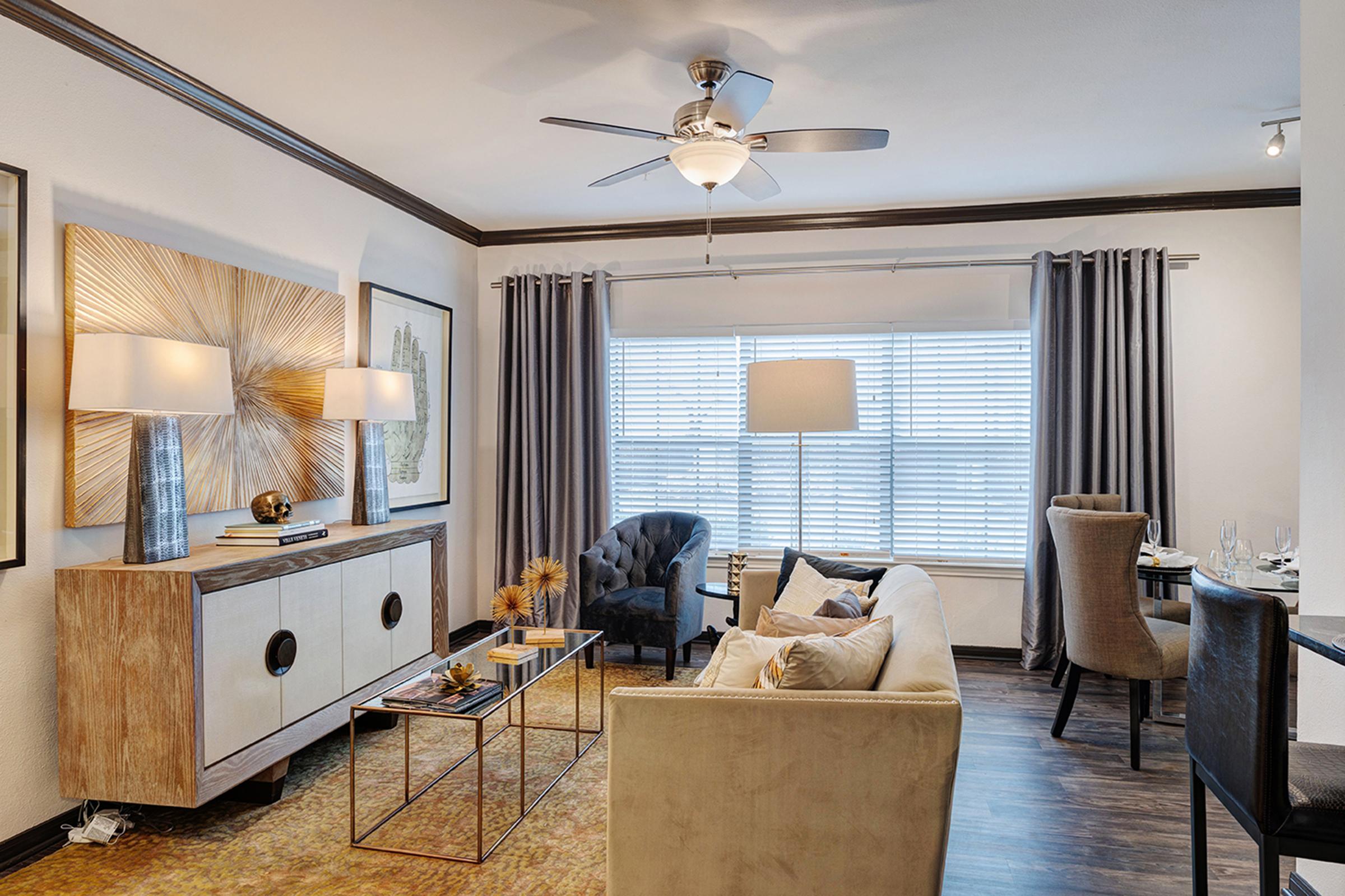 A modern living room featuring a beige sofa with patterned cushions and a decorative gold rug. There's a stylish sideboard, two elegant lamps, and a dark blue chair. Light streams in through a window adorned with grey curtains, and there's a dining area in the background with a round table and chairs.