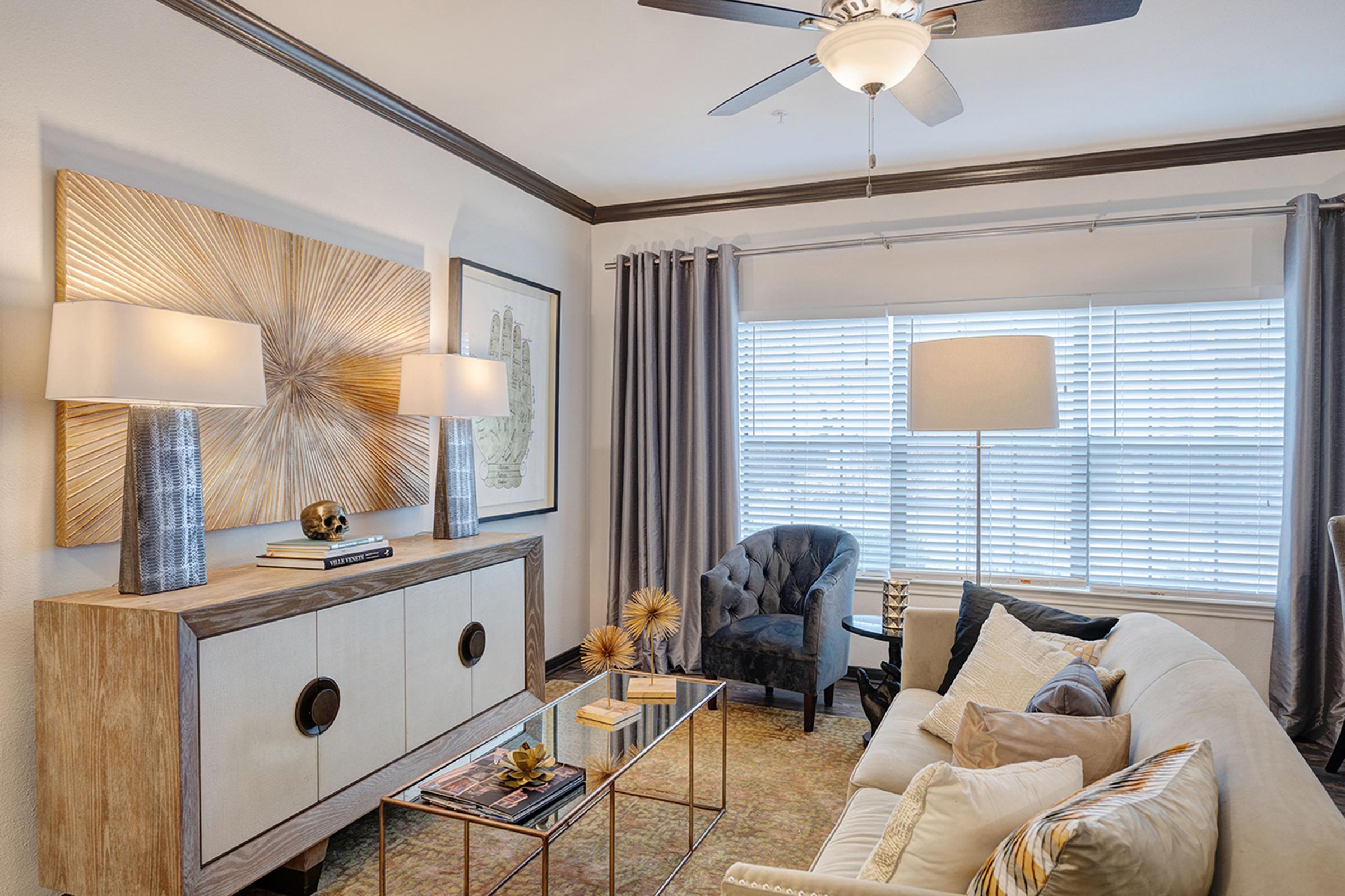A stylish living room featuring a beige sofa, a glass coffee table with decorative items, modern lamps on a sideboard, and a blue armchair. The walls are adorned with artwork, and large windows with blinds allow natural light to illuminate the space, complemented by neutral curtains and a warm color palette.