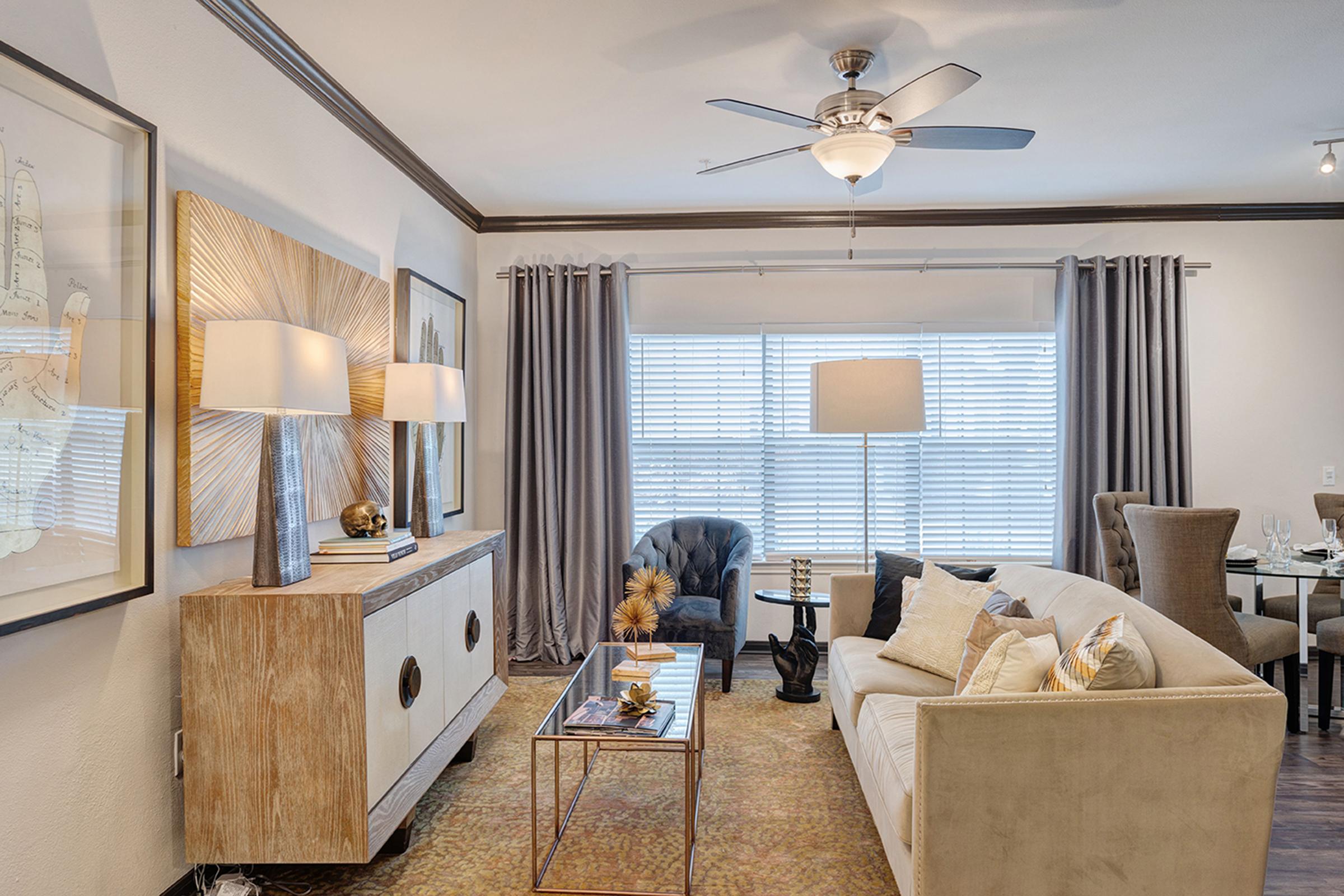 A modern living room featuring a cream-colored sofa with decorative pillows, a glass coffee table, and an elegant sideboard with lamps. There is a blue armchair, large windows with light gray curtains, and framed artwork on the walls. A dining area with chairs is visible in the background.