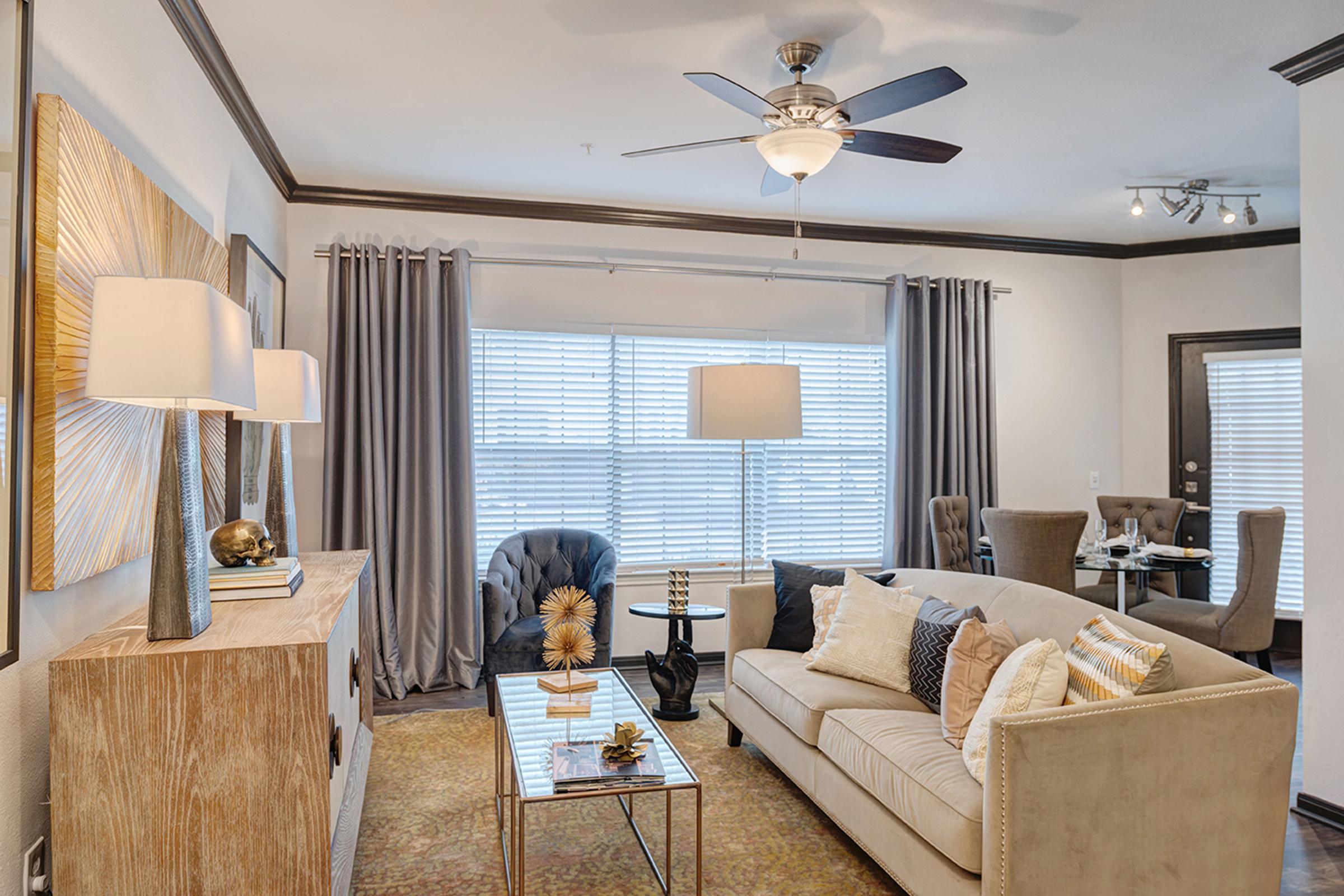 A modern living room featuring a beige sofa adorned with decorative pillows, a stylish coffee table, and a textured area rug. There are large windows with blinds, a ceiling fan, and contemporary decor, including wall art and table lamps. Two armchairs and a dining area are also visible, creating an inviting atmosphere.