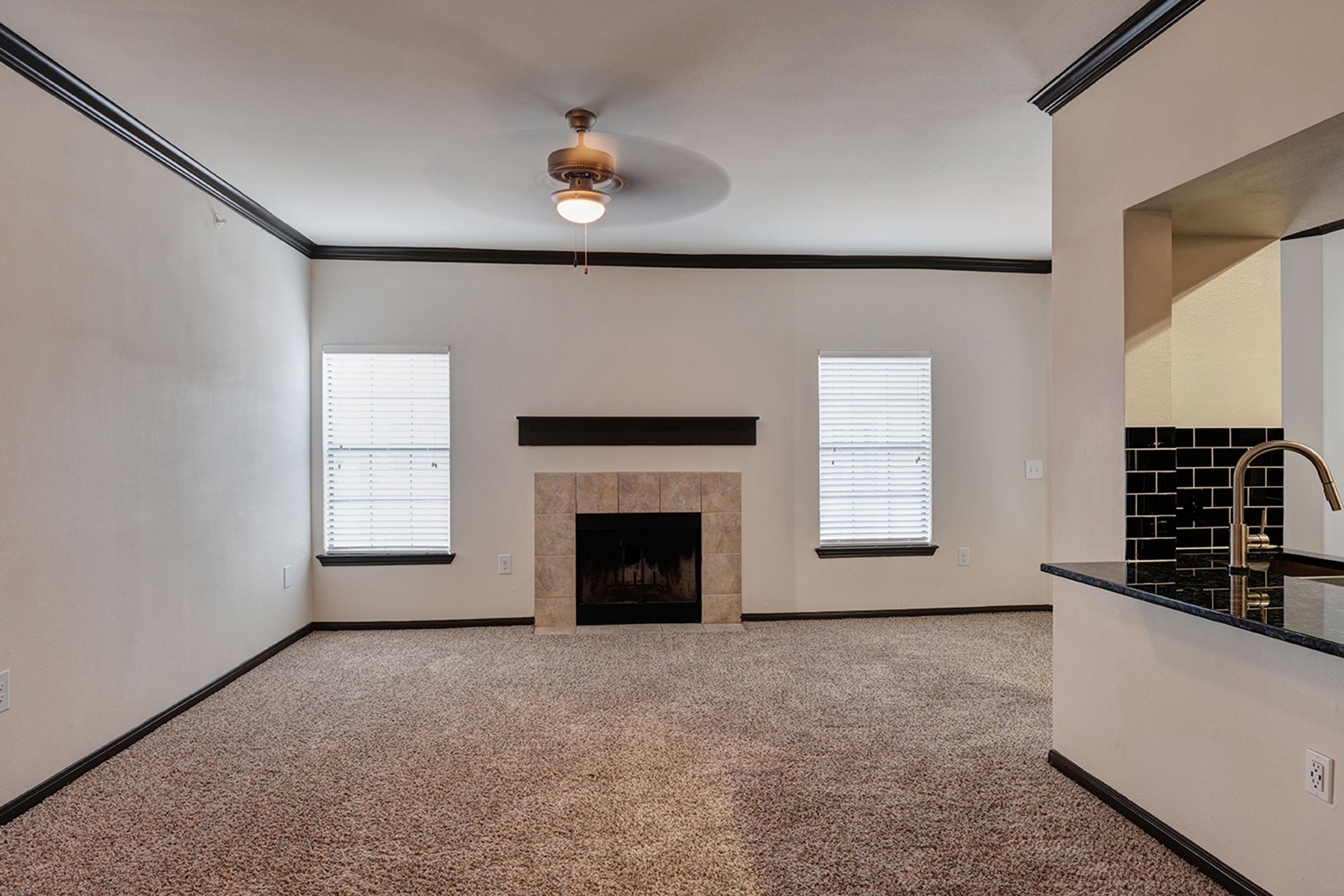 A spacious, well-lit living room featuring light-colored walls, a carpeted floor, and two windows with blinds. There is a decorative fireplace centered on one wall and a ceiling fan above. The room also includes an open layout leading to a kitchen area with dark countertops and a contrasting backsplash.