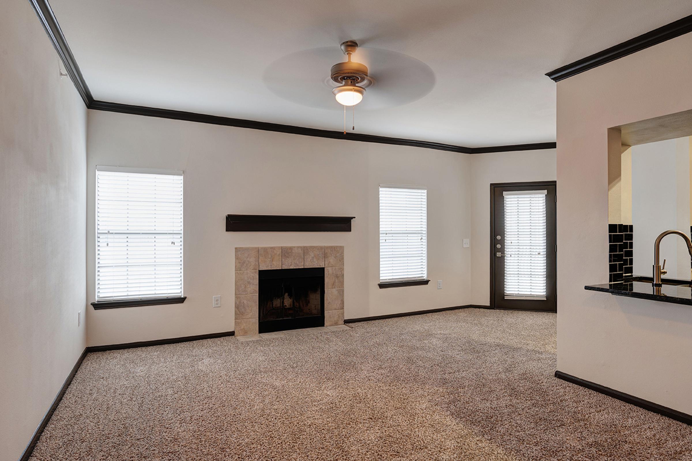 Spacious living room with light-colored walls and a ceiling fan. Features a decorative fireplace with a stone surround and two windows with blinds allowing natural light. The room has a cozy carpeted floor. A doorway leads to a kitchen area with dark cabinetry and a bar.