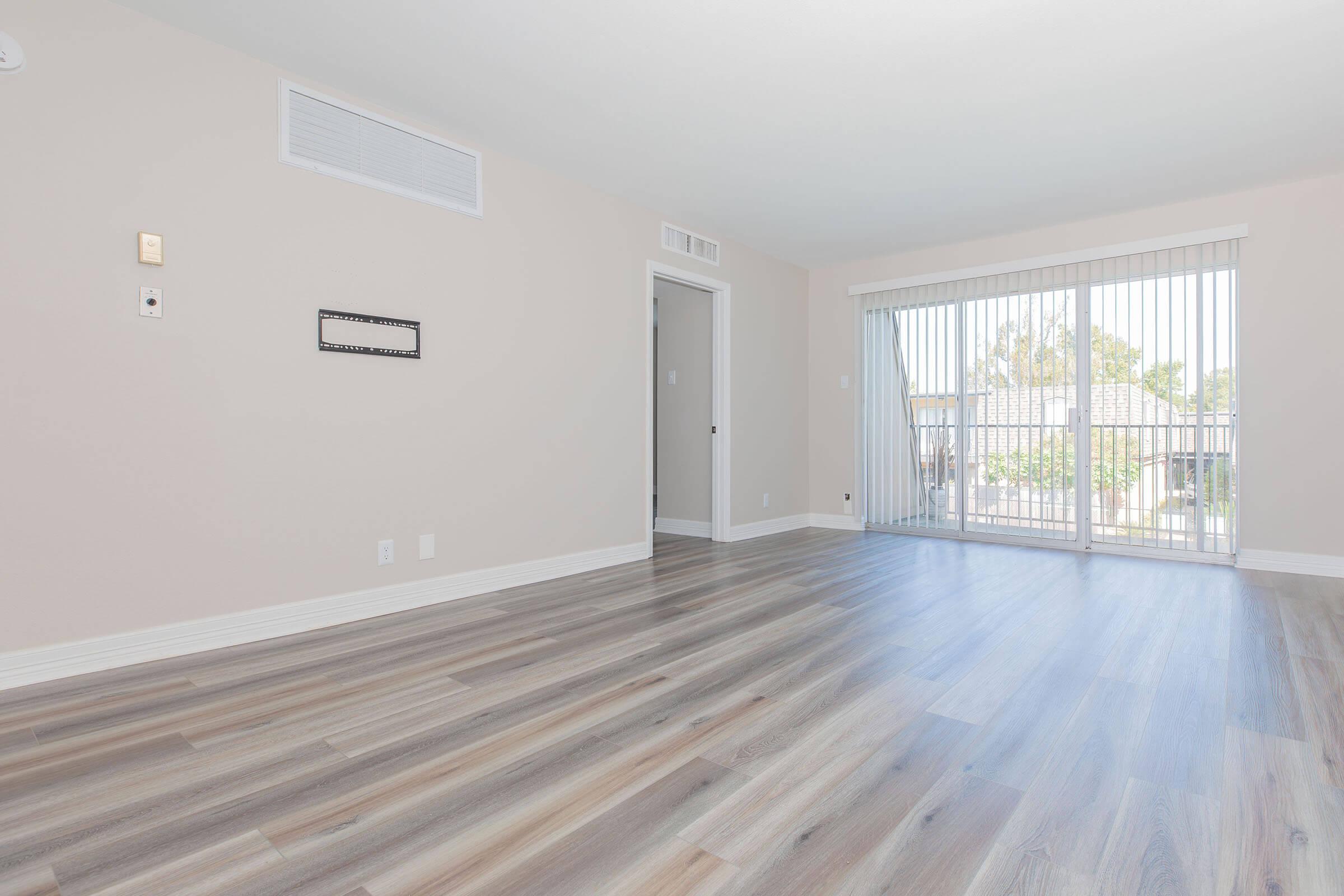 A spacious, vacant room with light-colored walls and hardwood flooring. There is a window with vertical blinds allowing natural light, and a door leading to another area. The room is minimally furnished, creating a clean and airy atmosphere.