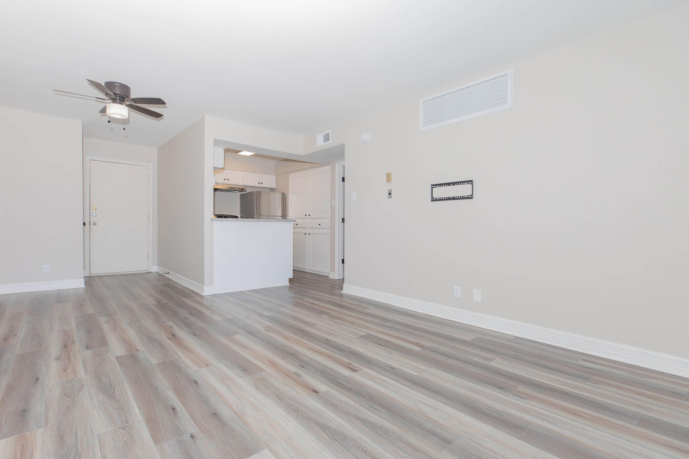 A spacious, modern living area with light-colored walls and hardwood-style flooring. The room features a ceiling fan, an open kitchen area with white cabinetry, and a doorway leading to an entrance. Natural light fills the space, creating a bright and inviting atmosphere.