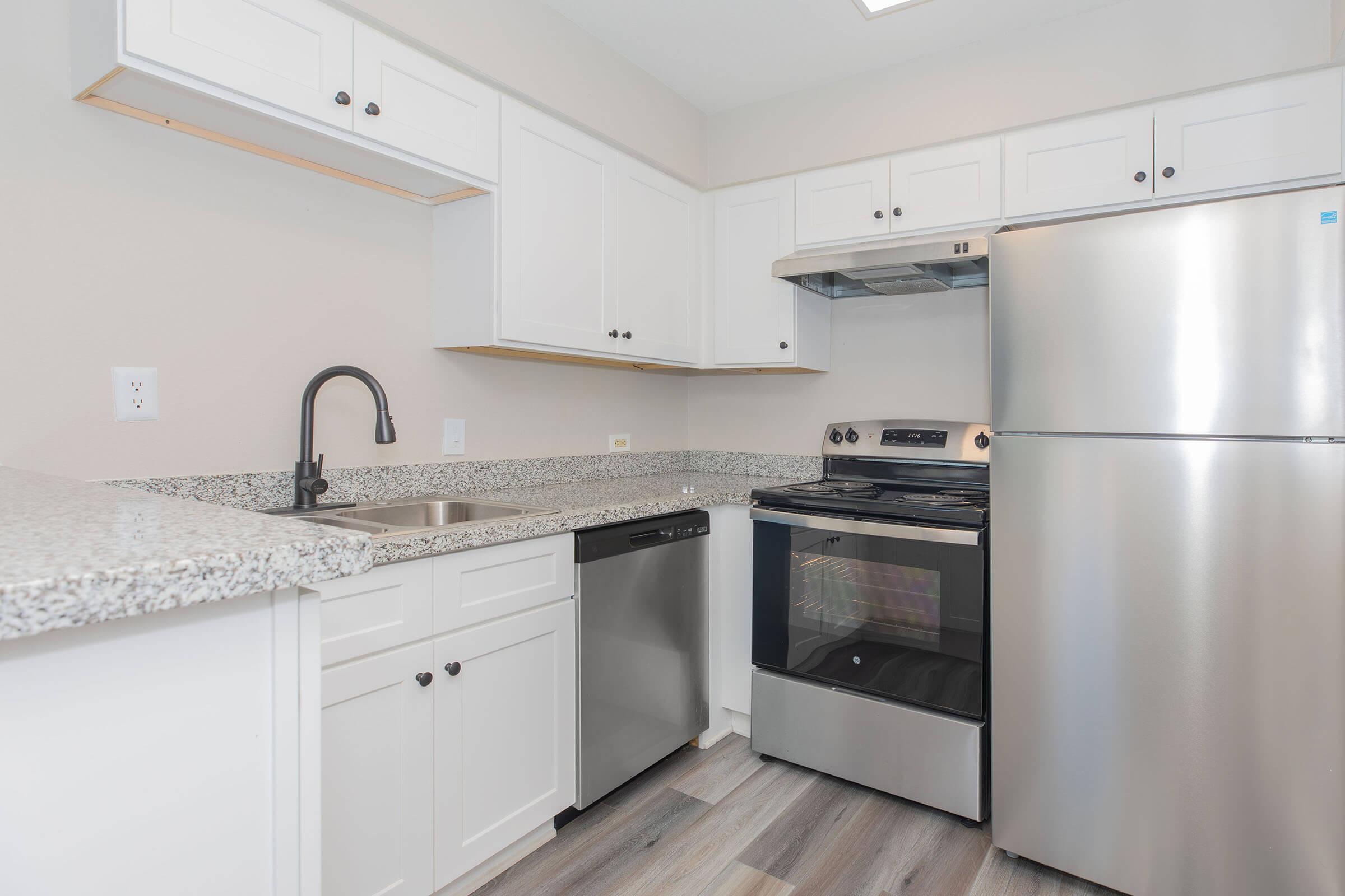 Modern kitchen featuring white cabinets, a stainless steel refrigerator, and a black oven and stove. The countertop is a light-colored granite, complemented by a black faucet over a stainless steel sink. The flooring is a light wood design. Bright, neutral color scheme throughout.