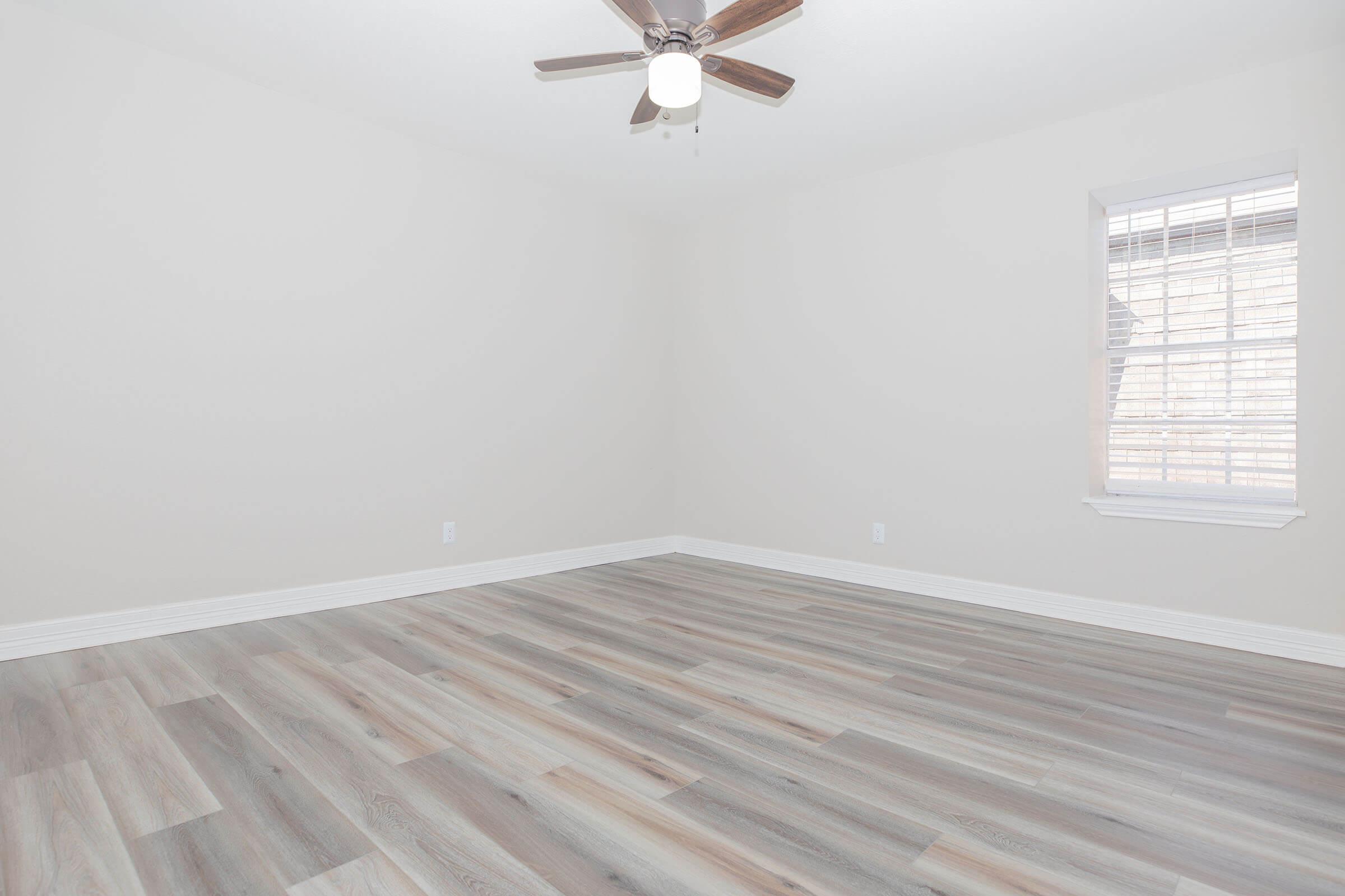 A vacant room with light-colored walls and a wooden floor. The ceiling features a ceiling fan with wooden blades. There is a window on one wall allowing natural light, and the space appears bright and spacious, ready for furnishing.