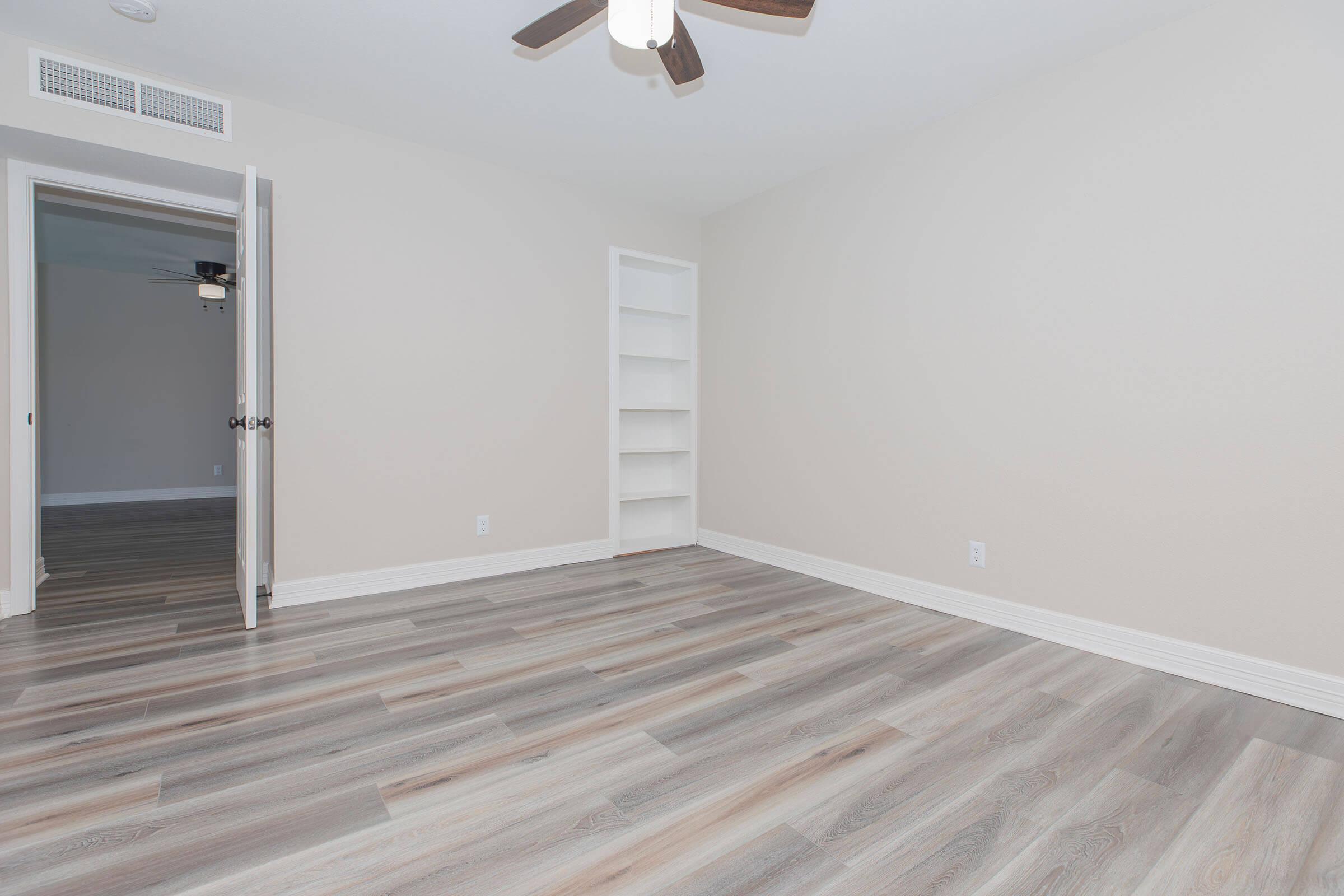 Empty room with light-colored walls and hardwood flooring. There is a ceiling fan, a door partially open leading to another room, and a built-in shelf against one wall. The space is well-lit and uncluttered, creating a bright and airy atmosphere.