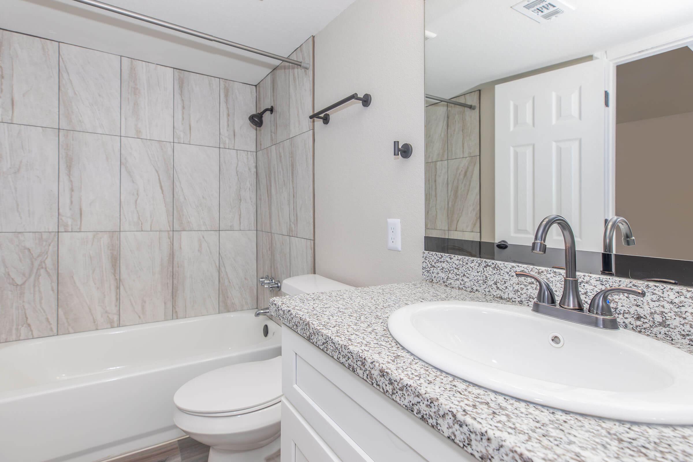 A modern bathroom featuring a white bathtub, a granite countertop sink, and a mirror. The walls are covered in light-colored tiles, and there is a towel bar mounted next to the mirror. A door leads to another room, enhancing the spacious feel of the area.