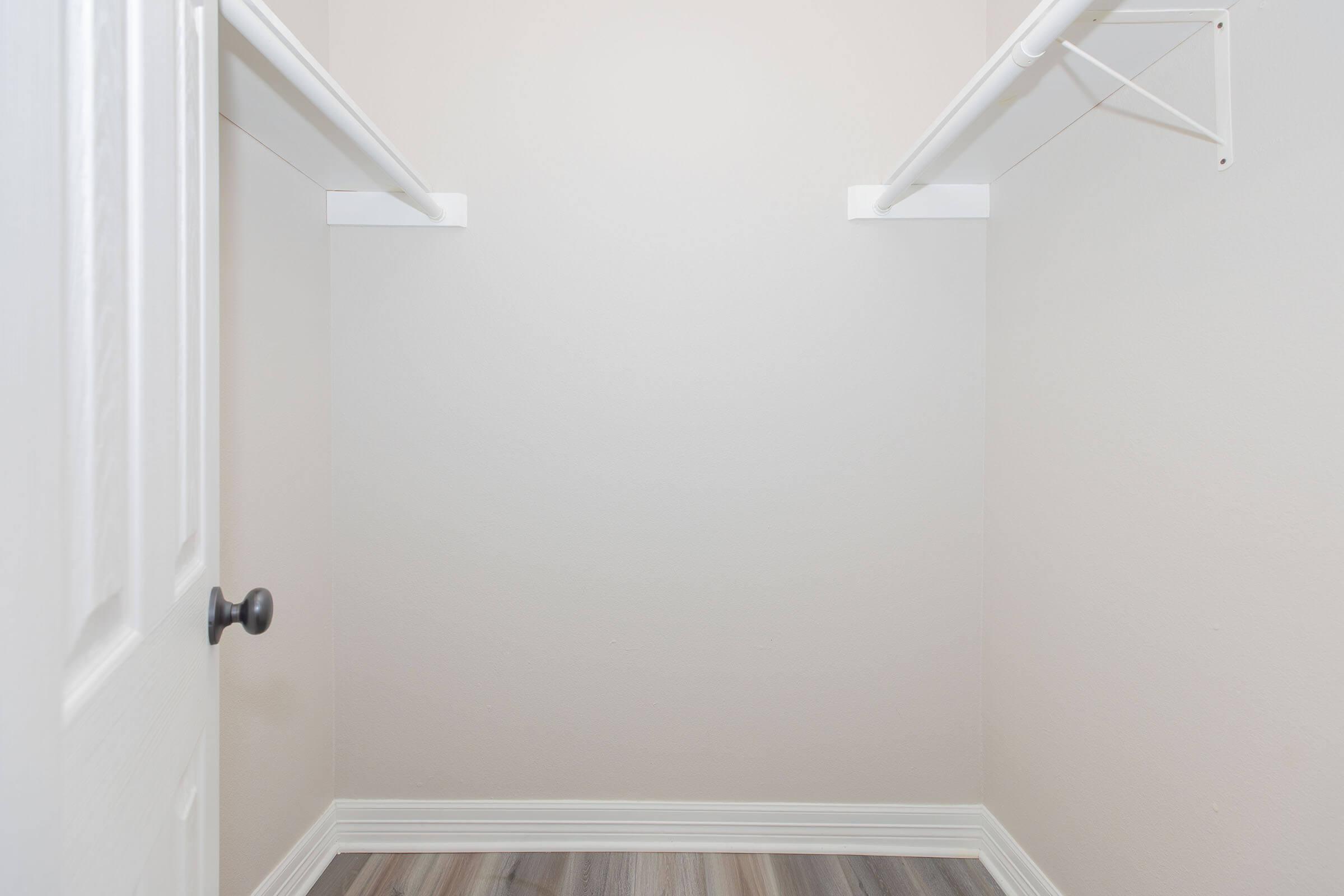 Empty closet with light-colored walls, featuring two white hanging rods. The floor is covered in laminate with a wood-like appearance. A closed door with a black knob is visible on the left side. The overall space appears spacious and uncluttered.