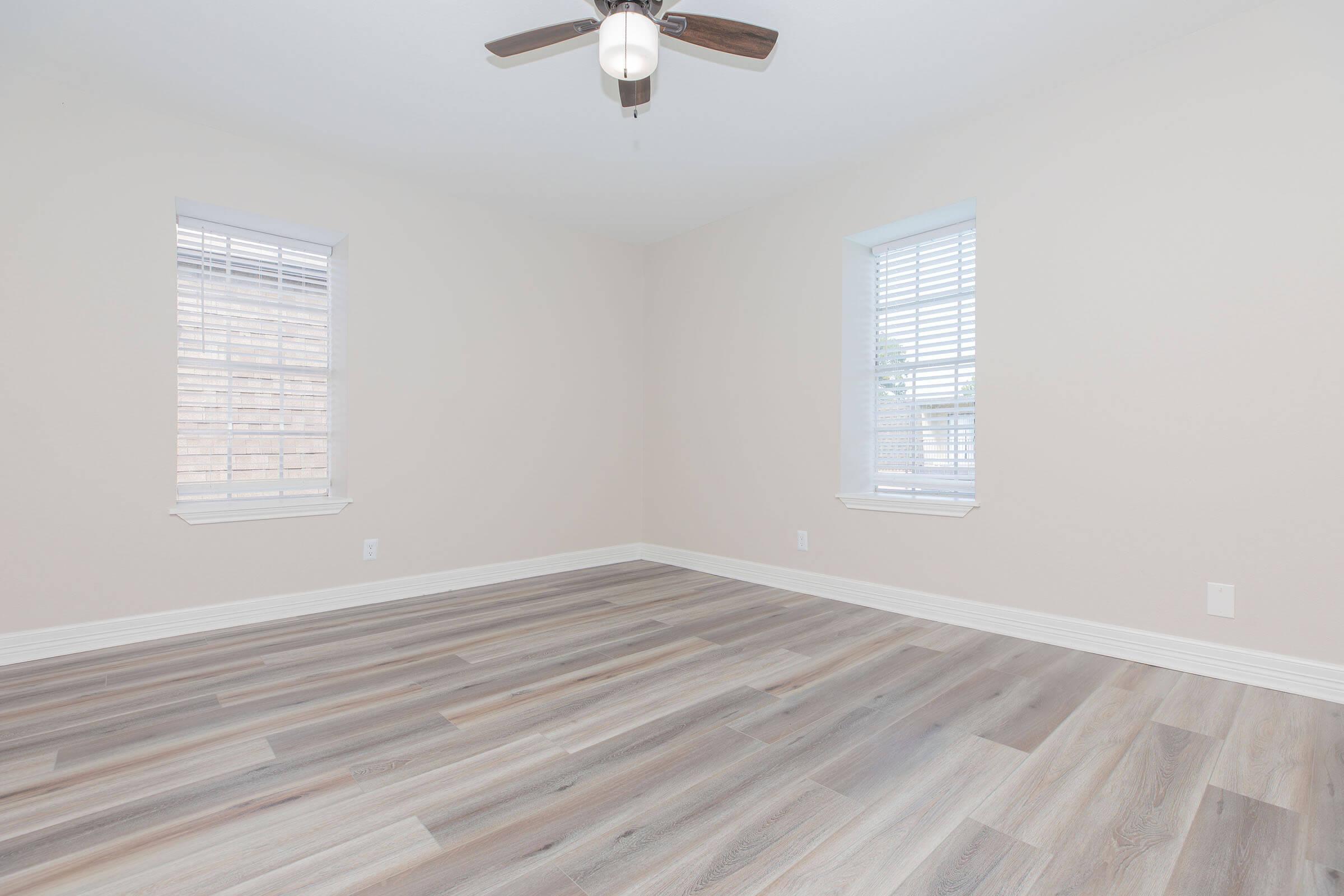 Empty room with light-colored walls, two windows with blinds, and a ceiling fan. The floor has a plank design in various shades of brown. The space is bright and spacious, suitable for various uses like a living area or bedroom.
