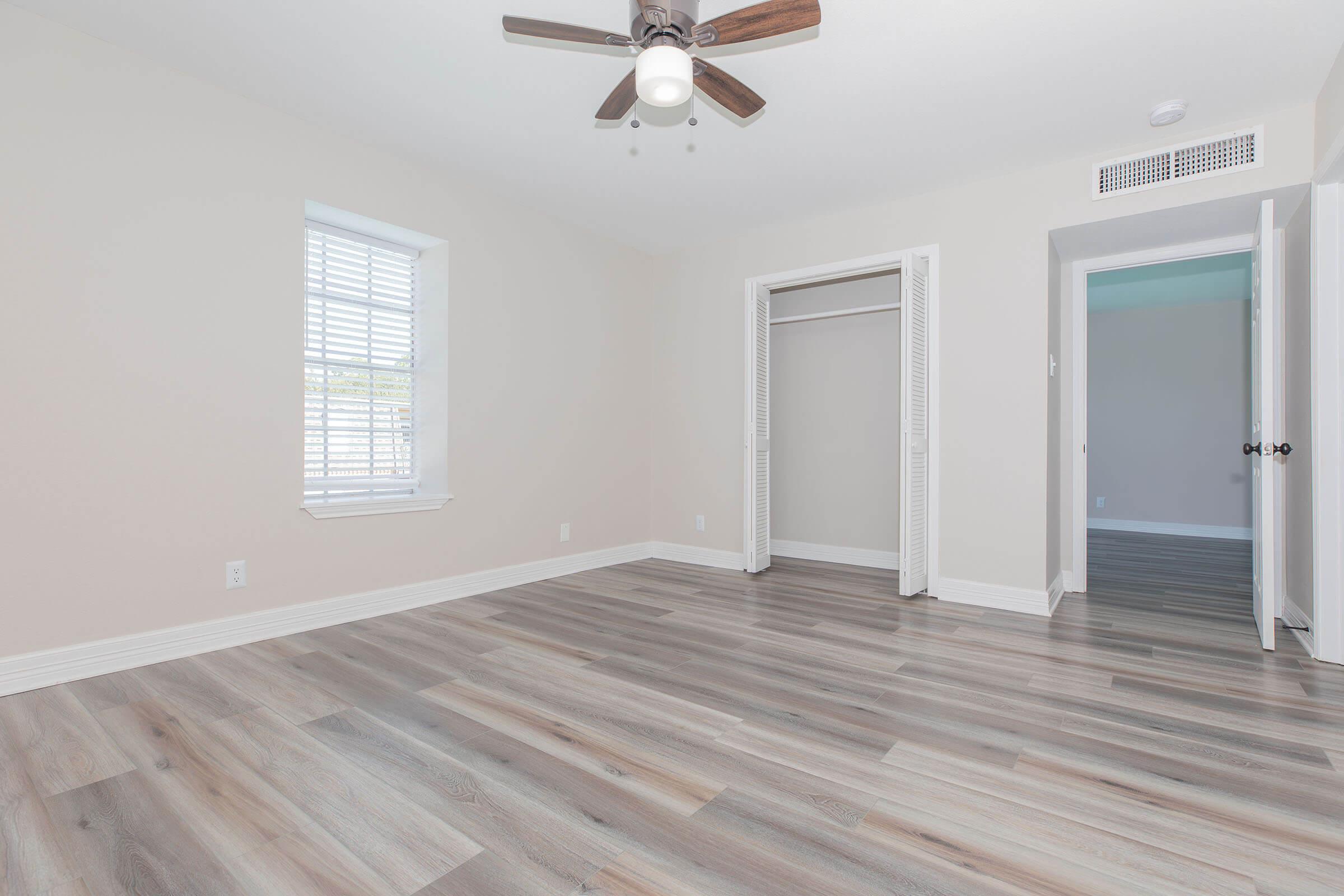 A clean, empty room featuring light-colored walls, a ceiling fan, and wooden laminate flooring. There is a window with white blinds and an open closet with sliding doors on one side, while a partially open door leads to another room, suggesting a bright and spacious atmosphere.