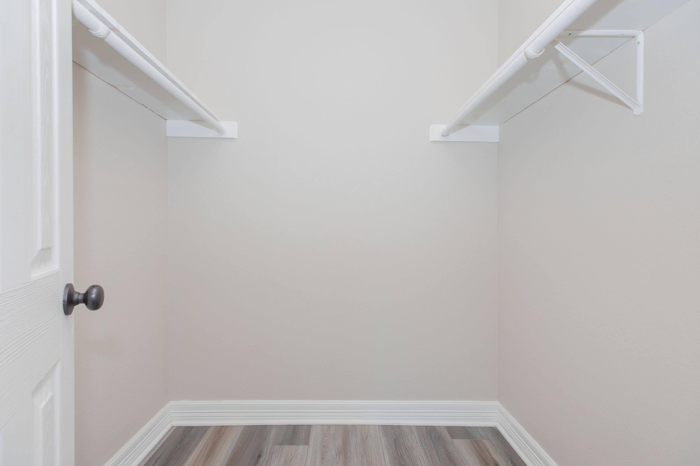 A tidy, empty closet with two white hanging rods against light gray walls, showcasing a clean and minimalist storage space. The floor features light gray and brown laminate planks, and there is a closed white door with a round black doorknob in the frame.
