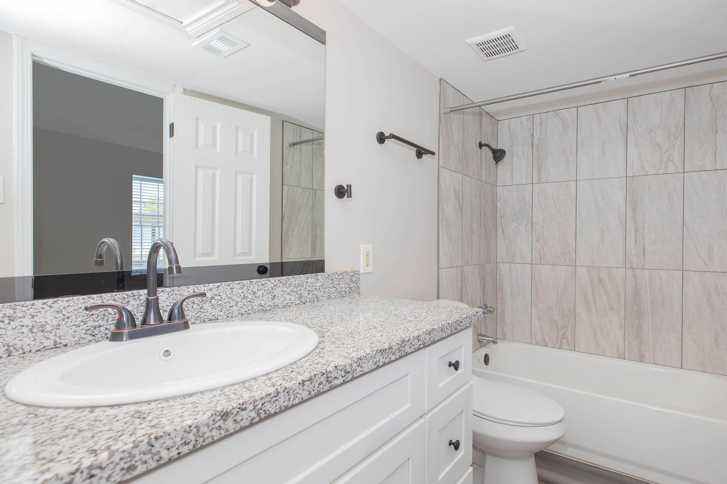 A modern bathroom featuring a white sink with a granite countertop, a bathtub with a shower, and light-colored tiled walls. There is a large mirror above the sink, a towel rack on the wall, and a door leading to another room. Natural light is visible through a window.