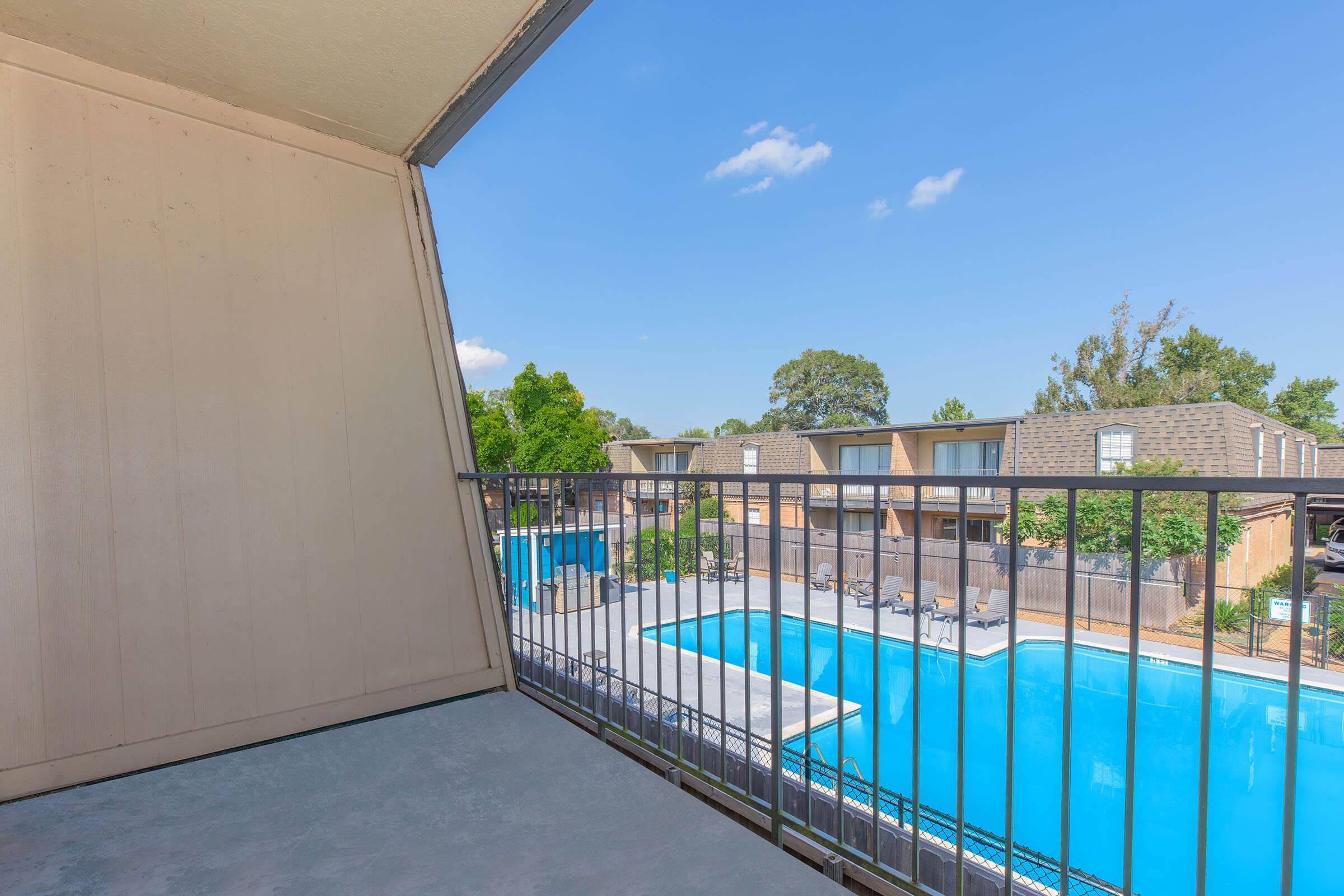 View from a balcony overlooking a clear blue swimming pool and surrounding apartment complex. The sky is bright and sunny with a few clouds, and the area is green with trees and landscaping. The pool area includes lounge chairs and is enclosed by a fence.