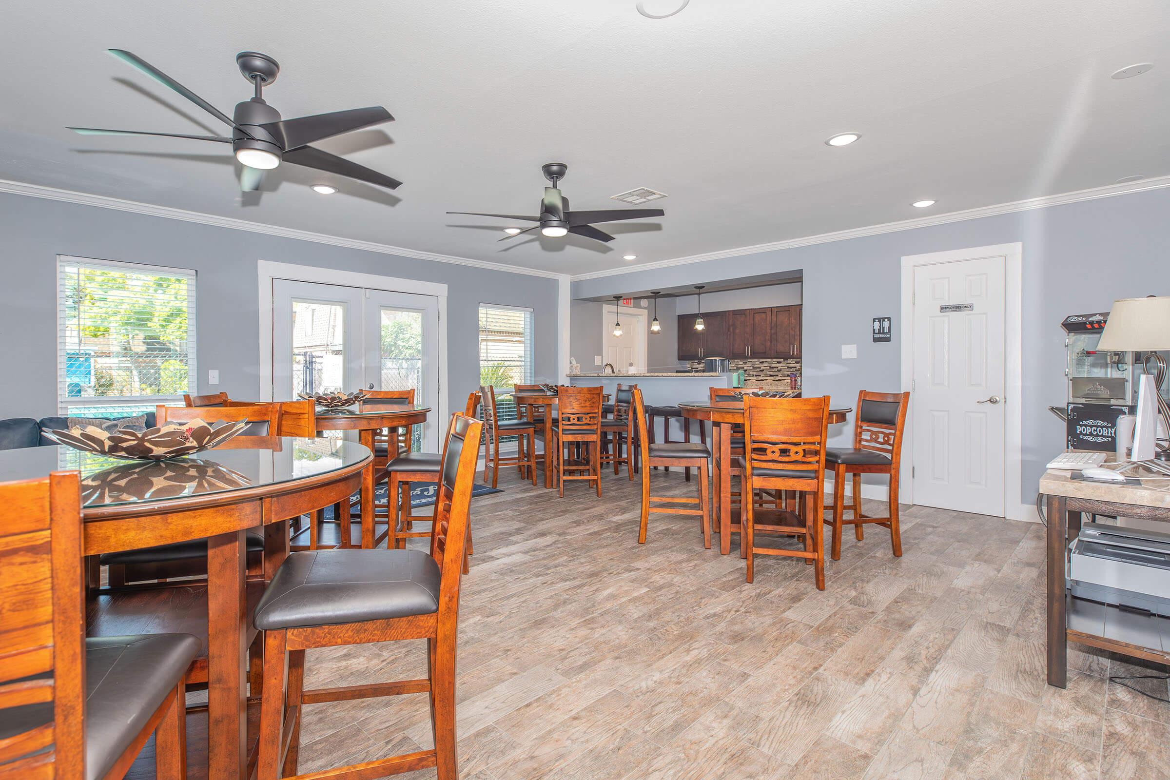 A spacious indoor dining area featuring wooden tables and chairs, elegant ceiling fans, and large windows allowing natural light. The decor is modern with a neutral color scheme, highlighting a welcoming ambiance. A kitchen area is visible in the background, enhancing the functionality of the space.