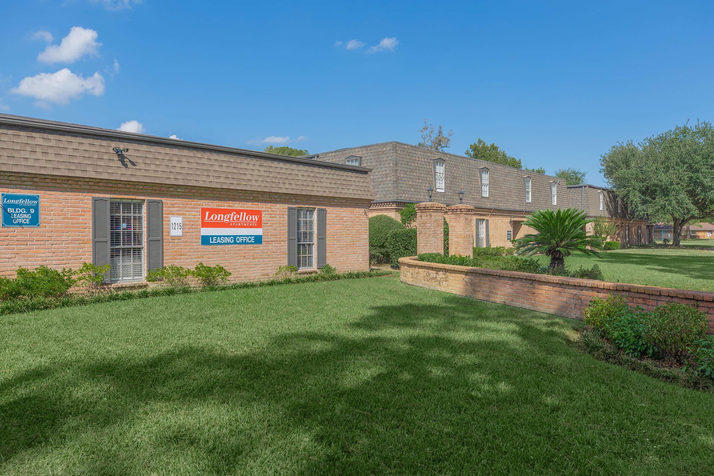 A leasing office of a residential complex, featuring a sign that reads "Longfellow Leasing Office." The building is made of brick with landscaped greenery and palm trees. The sky is clear with a few clouds, and the lawn is well-maintained.