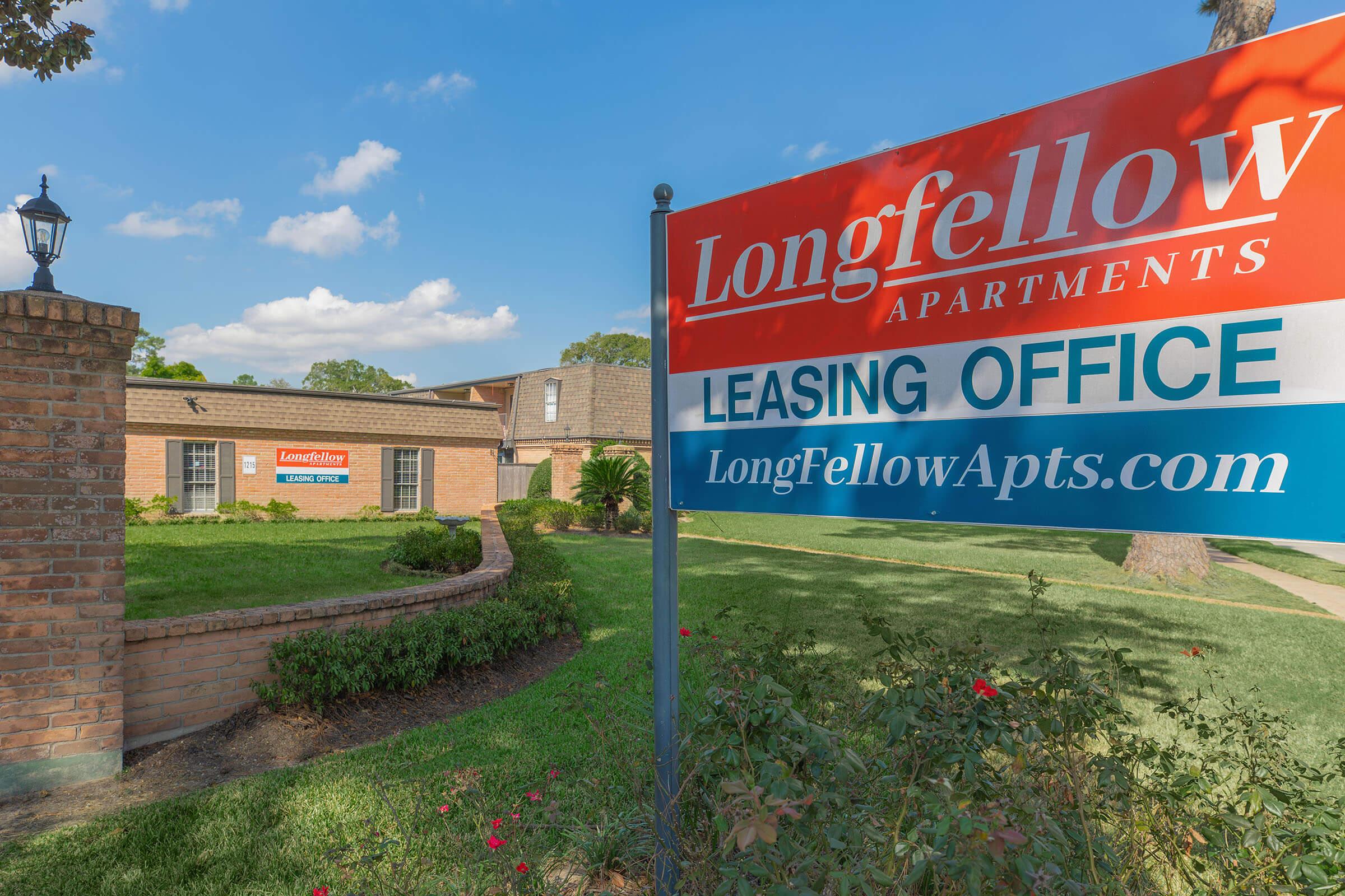 Sign for Longfellow Apartments with "Leasing Office" prominently displayed. The building features a well-maintained landscape with shrubs and grass. The sky is partly cloudy, contributing to a bright and inviting atmosphere. The website address for the apartments is also visible on the sign.