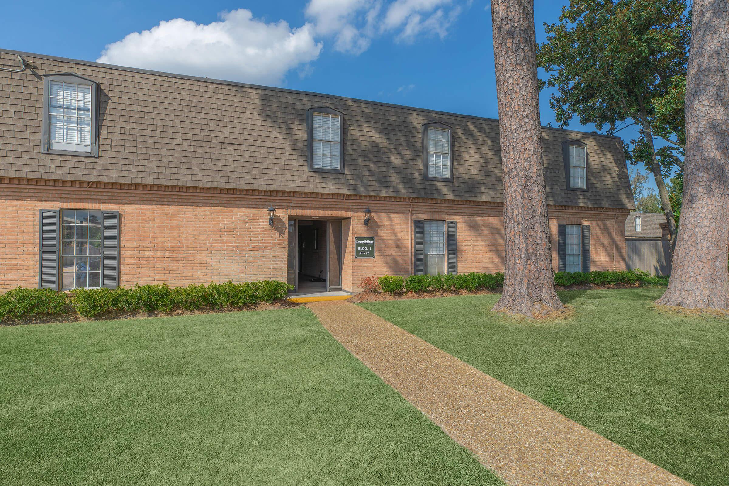 A brown brick building with a shingled roof and multiple windows on either side. The entrance features a pathway made of pebbles, bordered by well-maintained grass and shrubs. Tall trees stand on either side of the building under a clear blue sky.