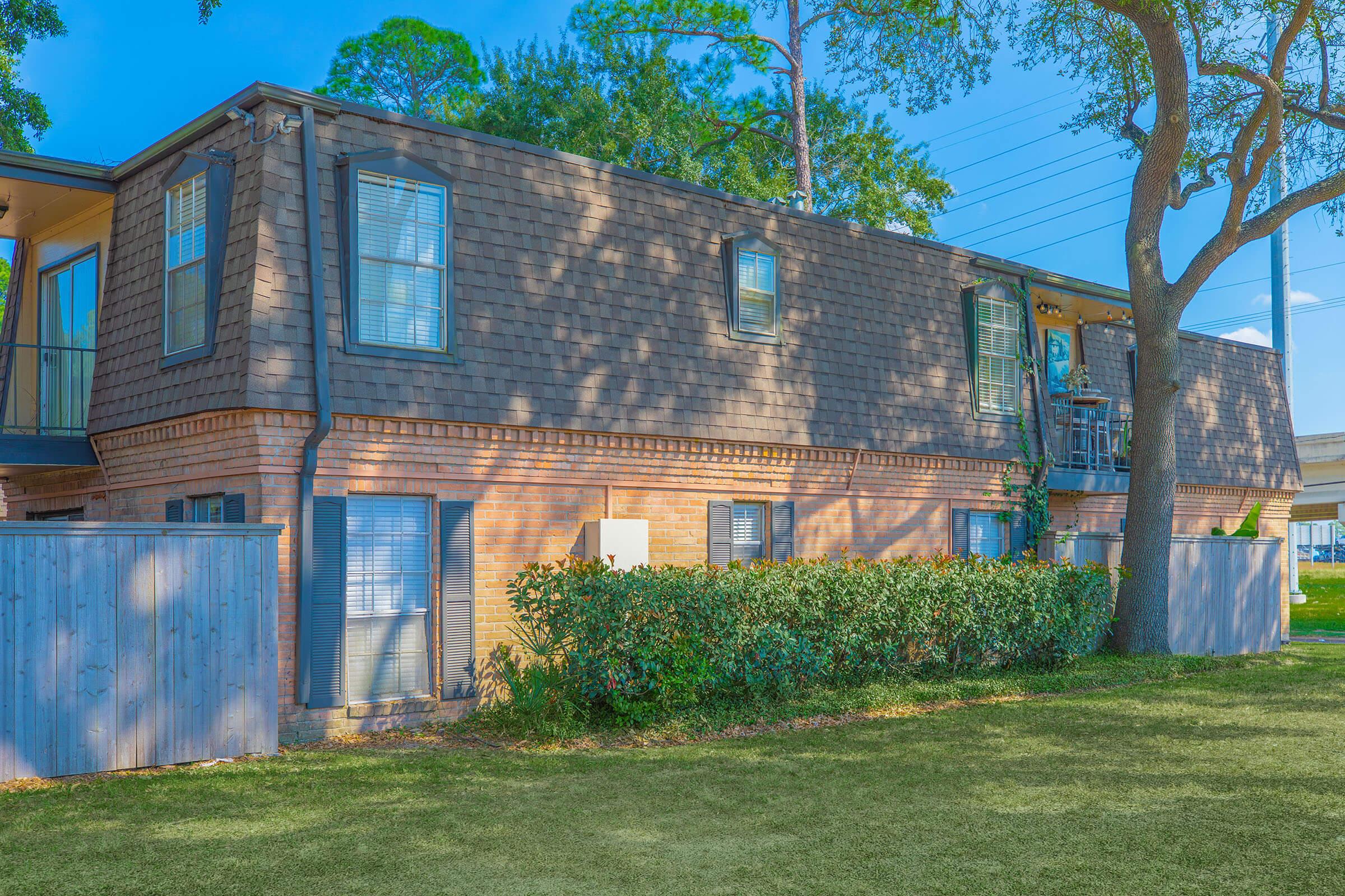 A two-story residential building with a brick facade and wooden siding, surrounded by greenery and grass. The building features multiple windows and a balcony on the upper level, with a fence and tree in the foreground. Bright blue sky and sunlight create a cheerful atmosphere.