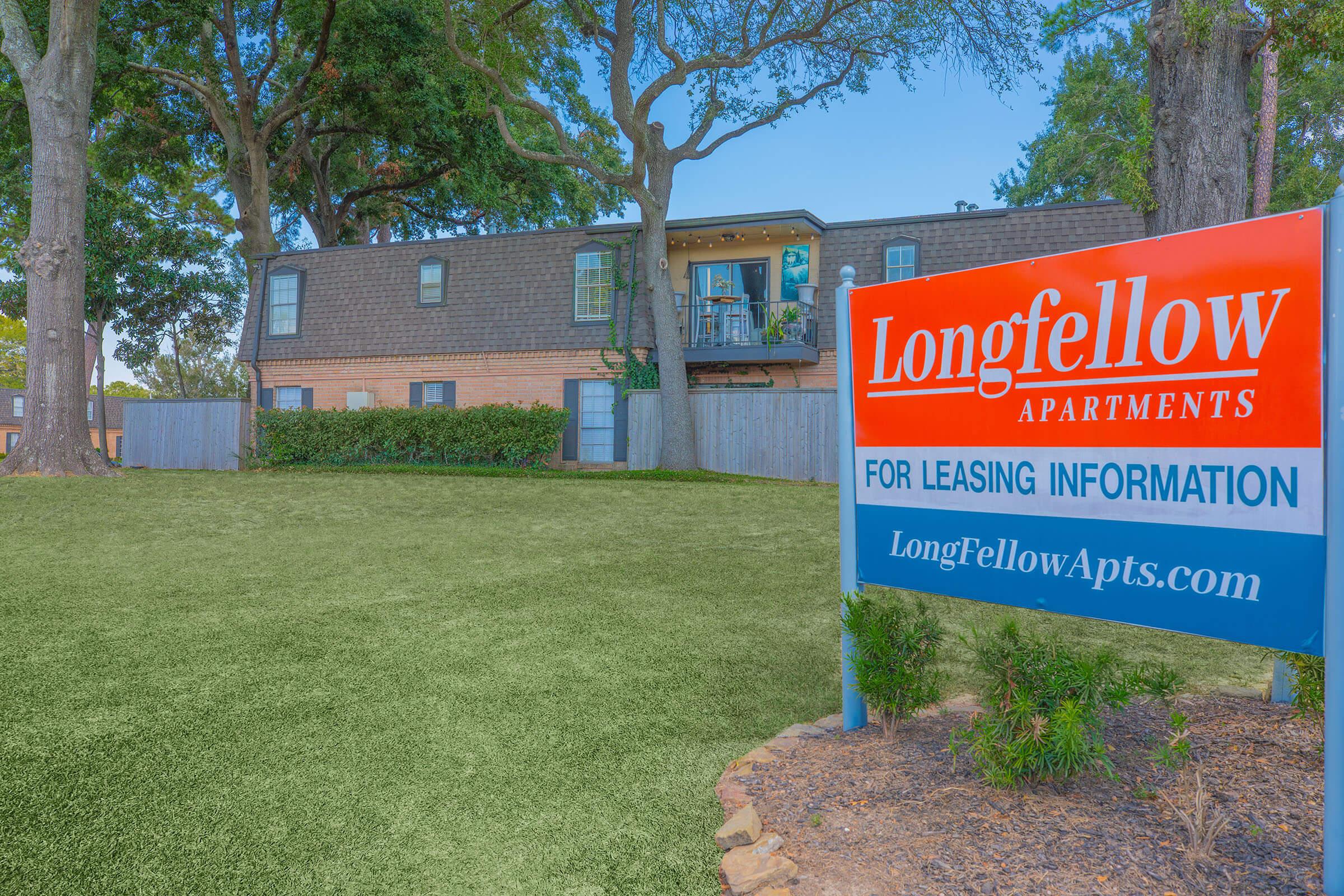 Sign for Longfellow Apartments with leasing information, set in front of a two-story building surrounded by trees and green grass. The apartment complex appears inviting, featuring a balcony on the upper level and a well-maintained lawn.