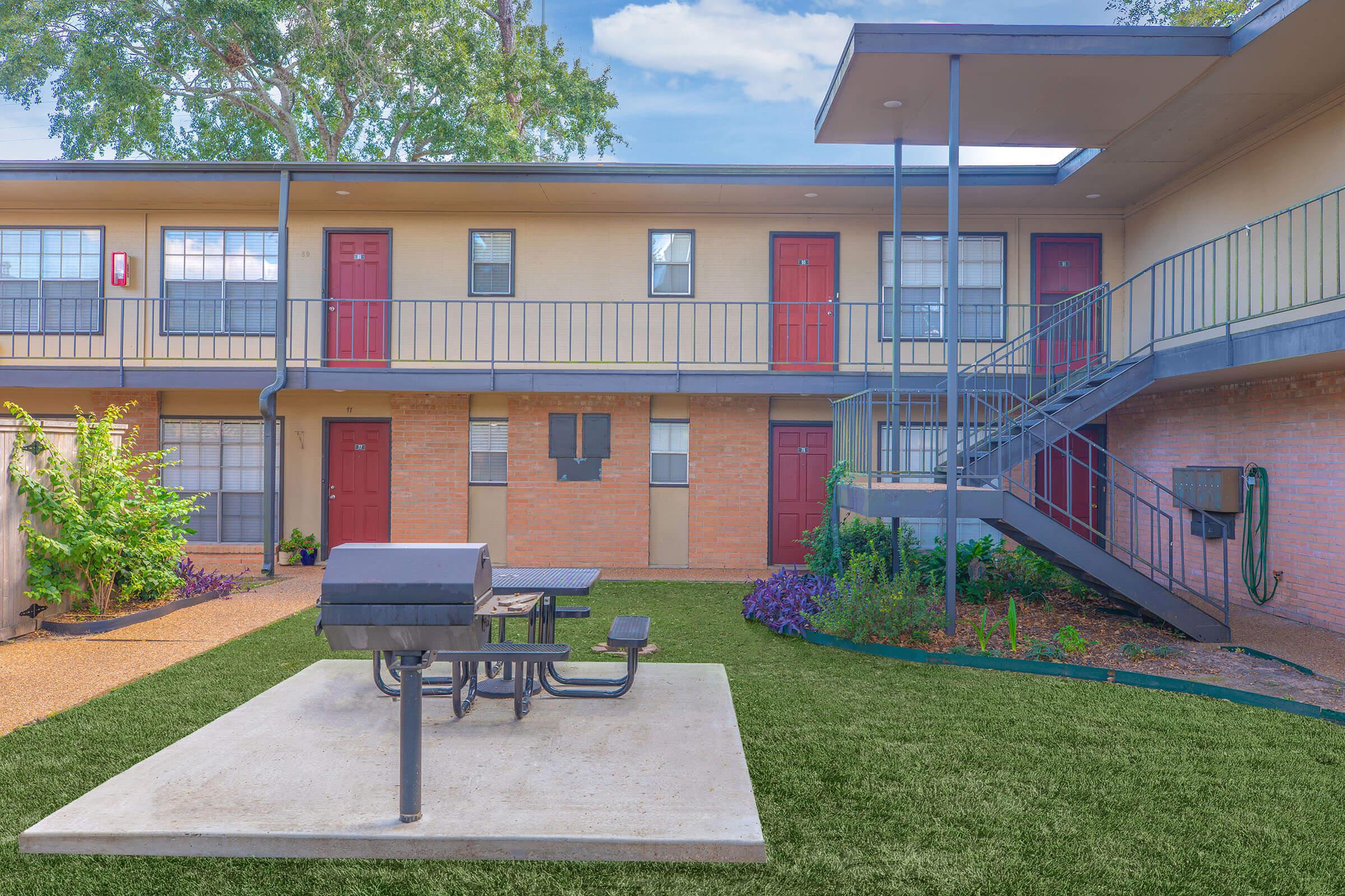 An outdoor area of an apartment complex featuring a concrete picnic table and grill, surrounded by green grass and colorful plants. In the background, there are two levels of apartments with red doors and a staircase leading to the upper floor. The scene is bright and inviting, under a clear blue sky.