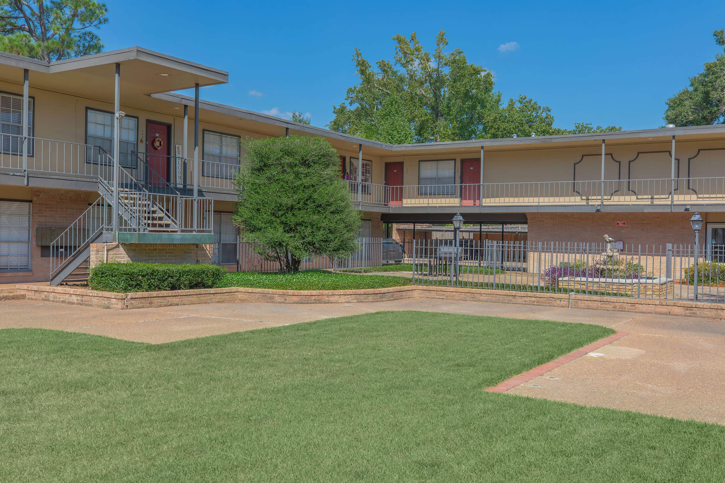 A two-story apartment complex with a well-maintained courtyard. The building features balconies, stairs, and a landscaped area with shrubs. The sky is clear and blue, and the pathway is made of stone.