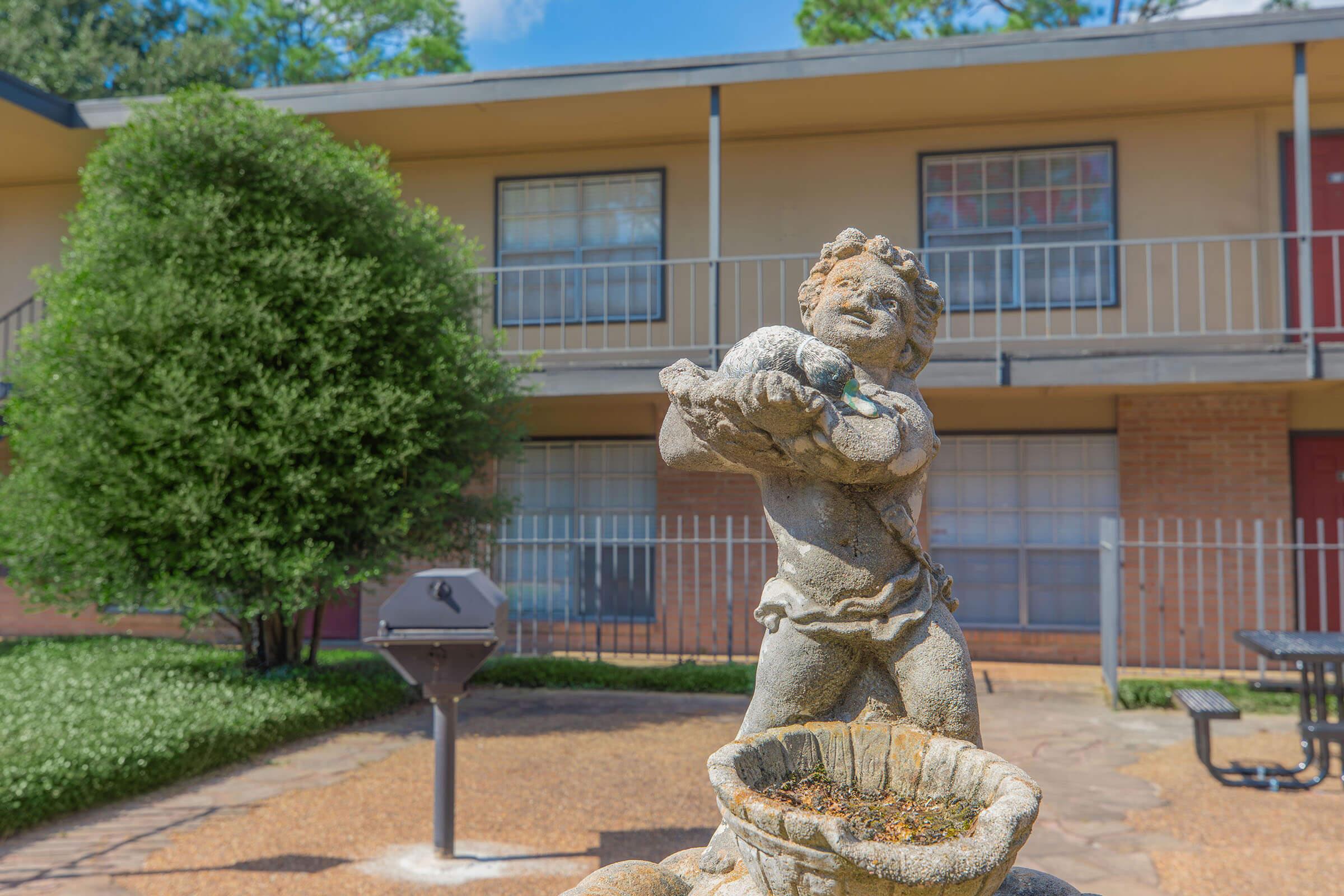A stone statue of a cherubic figure holding a bird, positioned in a landscaped courtyard featuring green grass and a background of a two-story building with balconies. A barbecue grill and picnic table are visible nearby under blue sky with scattered clouds.