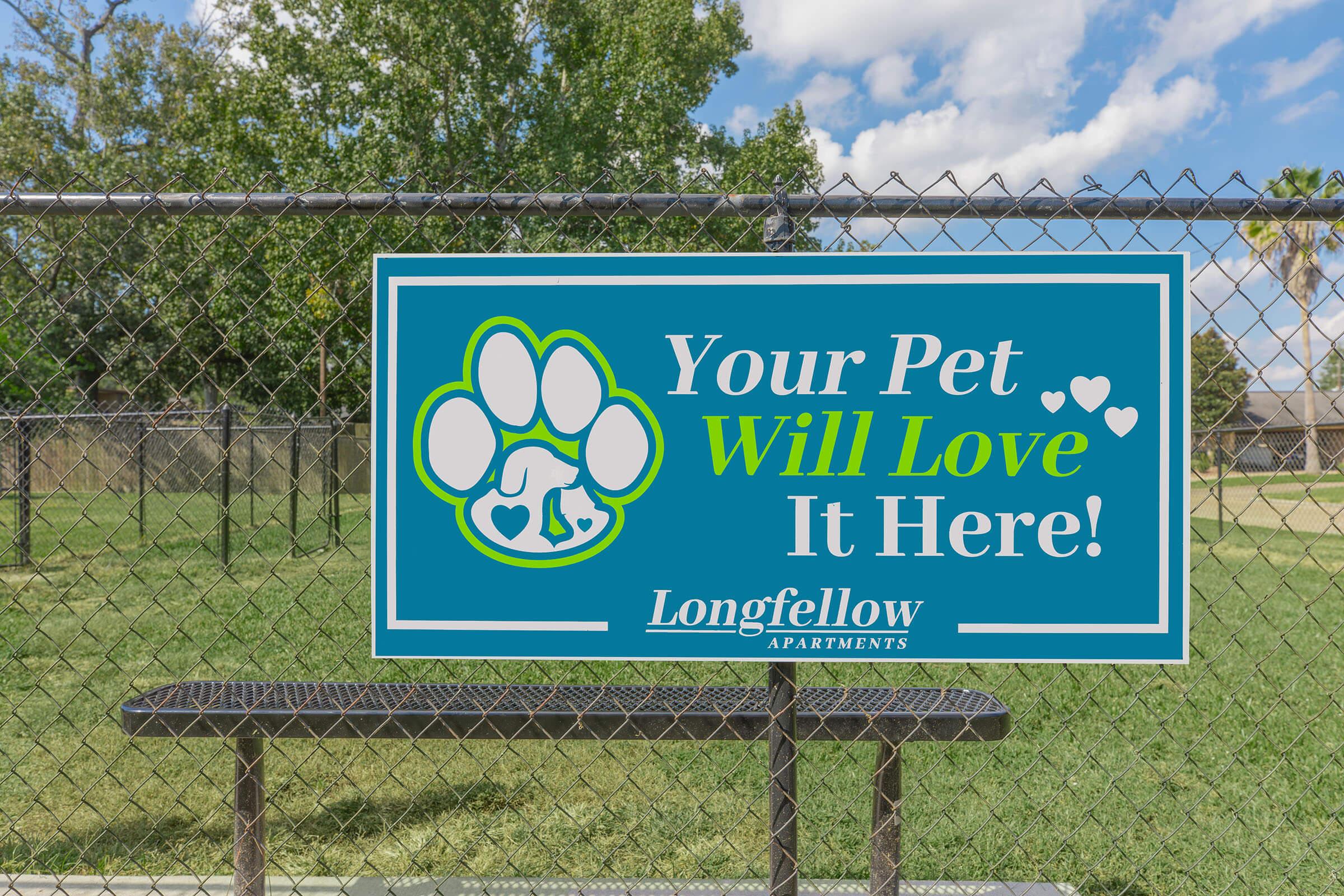 A colorful sign at a pet-friendly area features a paw print graphic and reads: "Your Pet Will Love It Here!" The sign is displayed on a green field with a chain-link fence in the background, promoting Longfellow Apartments as a welcoming place for pets.