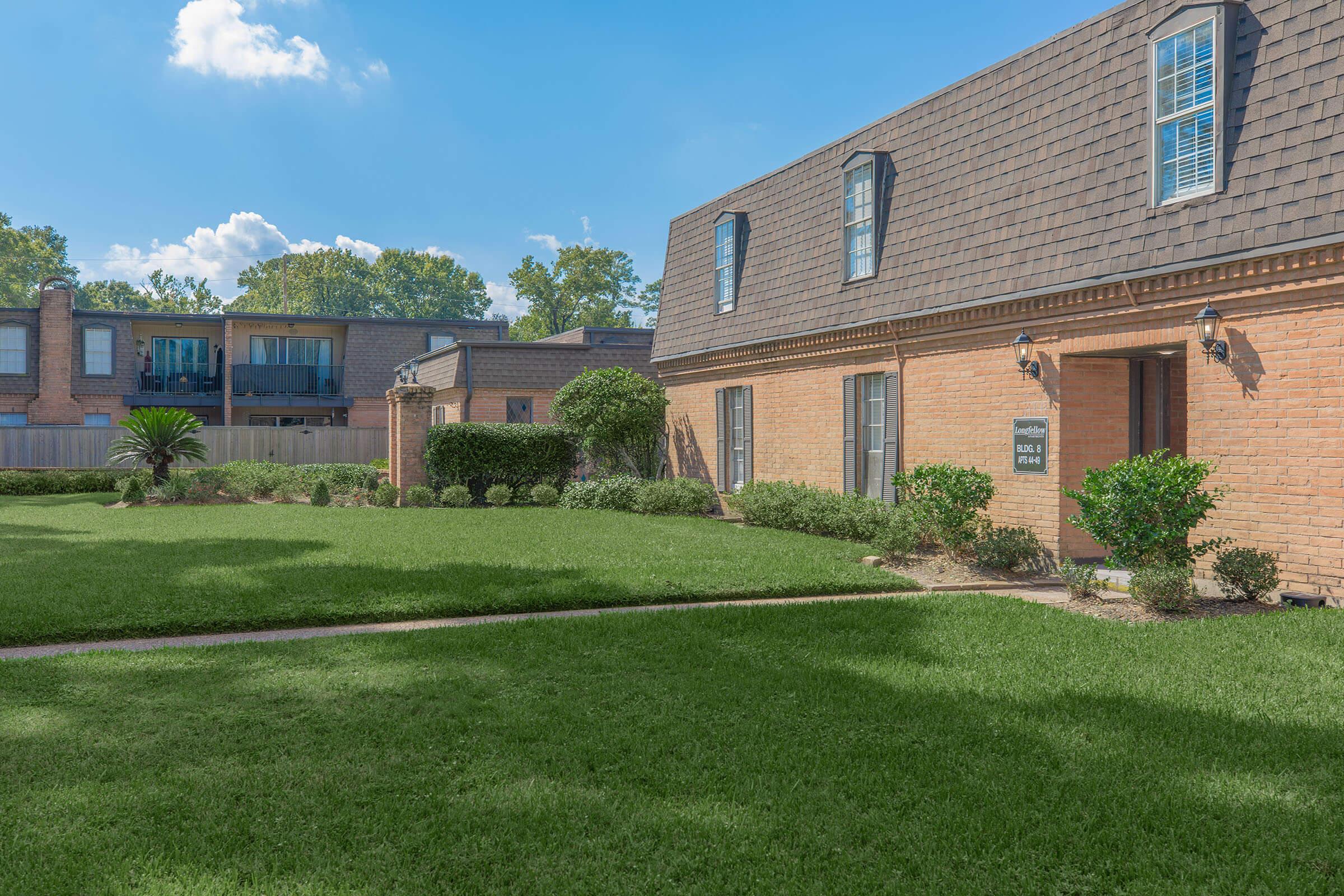 A well-maintained residential building with brick exterior, featuring several windows and a landscaped lawn. The surrounding area includes well-trimmed hedges and a pathway leading to the entrance. In the background, another apartment building is visible, surrounded by green trees under a clear blue sky.
