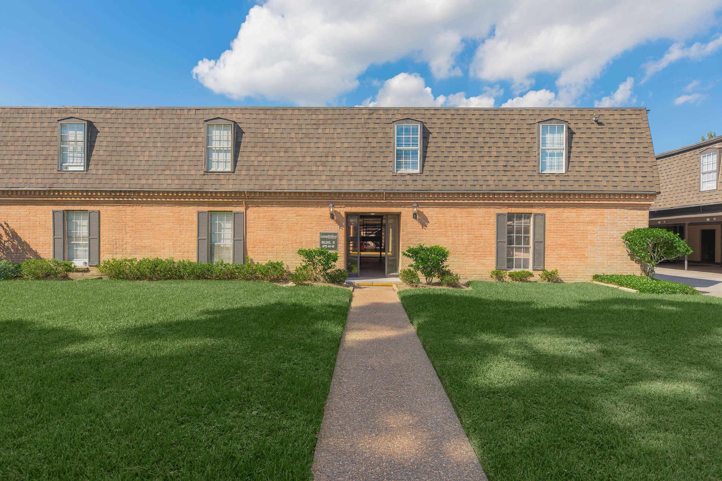 Brick building with a sloped roof, featuring multiple windows and a pathway leading to the entrance. The well-manicured lawn is green, with shrubs on either side of the path. The sky is partly cloudy, creating a pleasant outdoor scene.