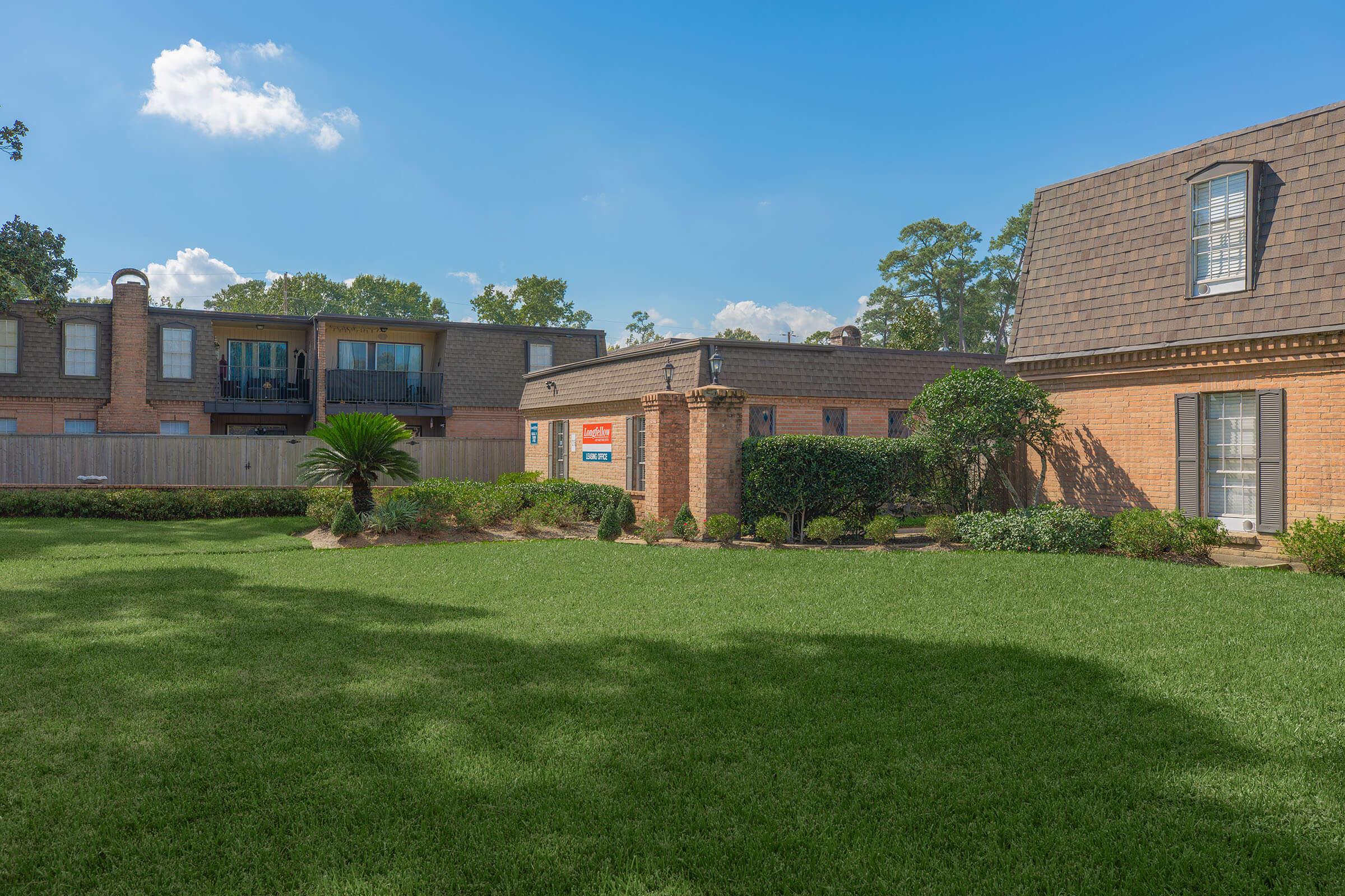 A spacious green lawn in the foreground with well-maintained shrubs. In the background, a two-story brick building and another building with balconies are visible. The sky is clear with a few clouds. A “For Sale” sign is placed near the entrance of one of the buildings.