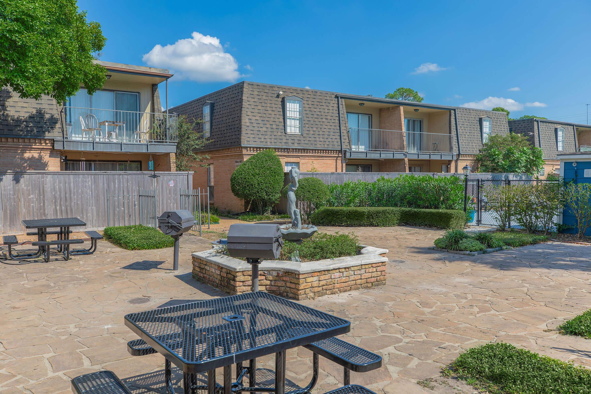 A landscaped courtyard featuring a stone barbecue grill and picnic tables. Surrounding the area are well-maintained bushes and a small statue. In the background, there are two apartment buildings with balconies and patio furniture. The sky is clear with a few clouds, creating a pleasant outdoor setting.