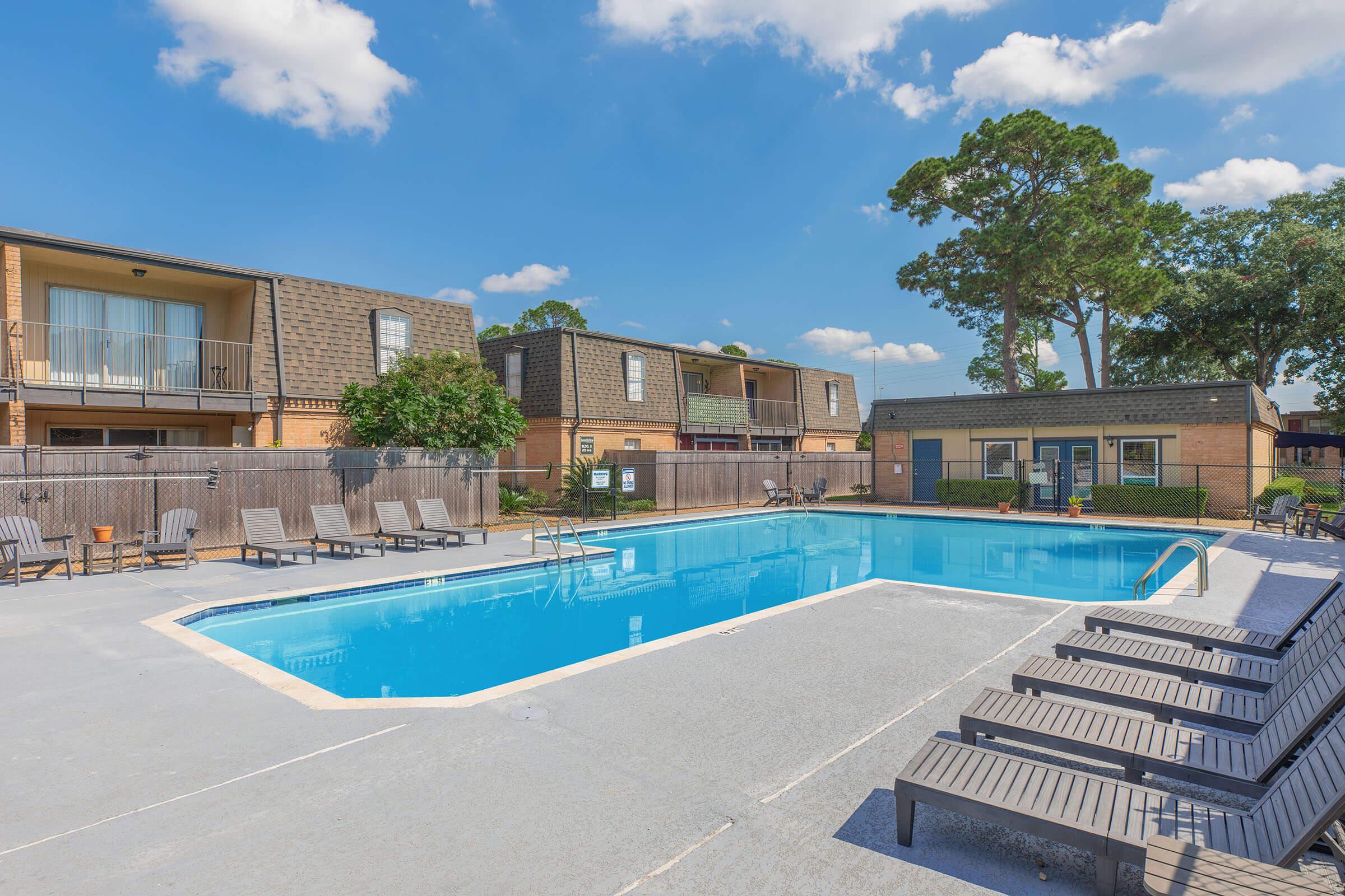 A clear blue swimming pool surrounded by lounge chairs, with apartment buildings and greenery in the background. The sky is bright with a few clouds, creating a warm and inviting atmosphere.